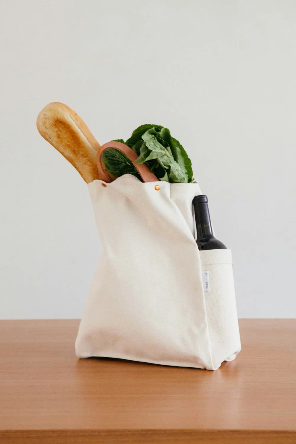 A white tote bag containing a baguette, lettuce, a sliced cucumber, and a bottle of wine on a wooden surface.