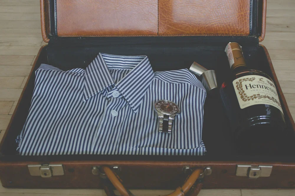 Open vintage suitcase containing a folded striped dress shirt, a watch, a flask, and a bottle of Hennessy cognac.