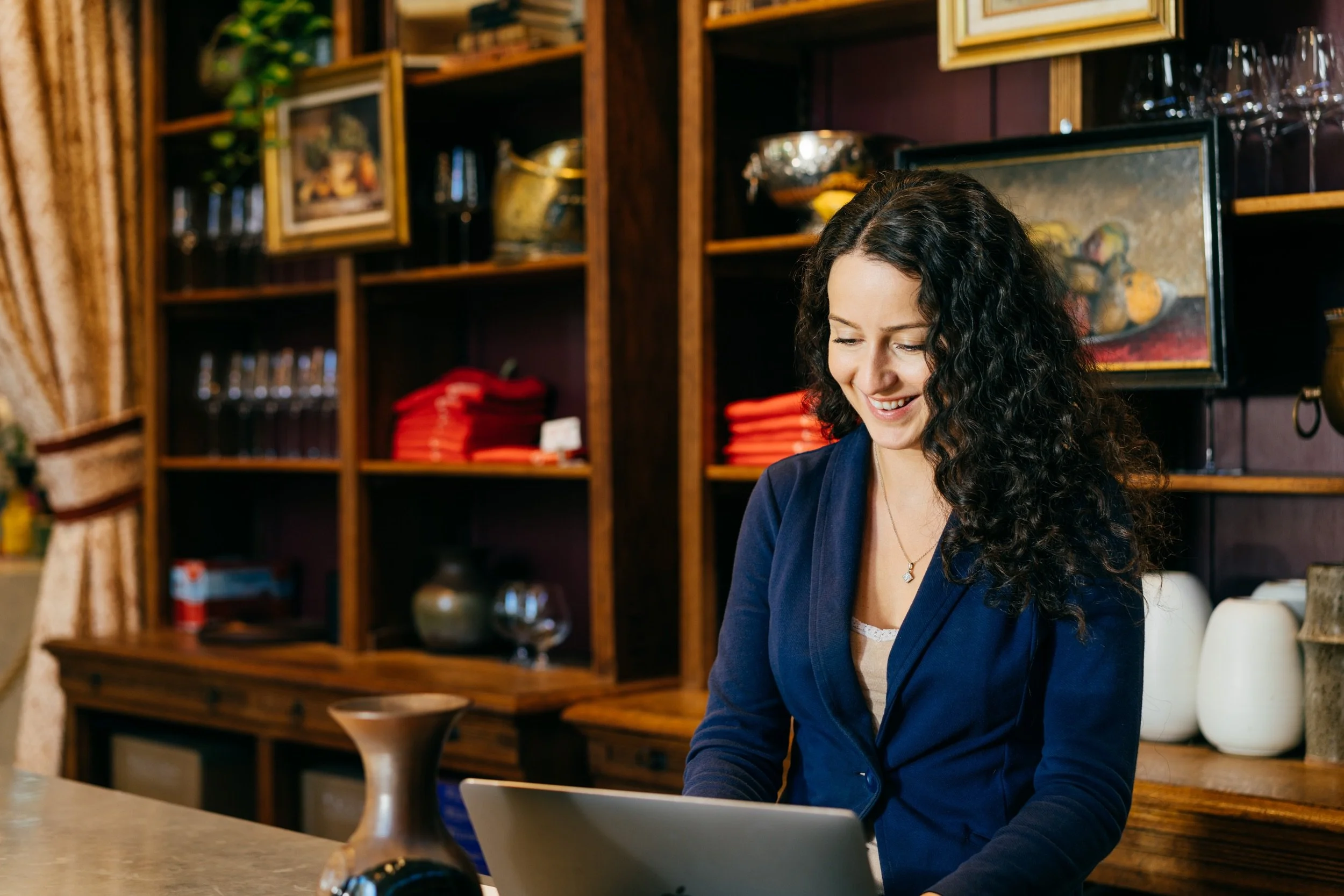 A woman with curly hair smiling and looking down at a laptop in a cozy, wood-paneled room filled with books, framed art, and decorative items.