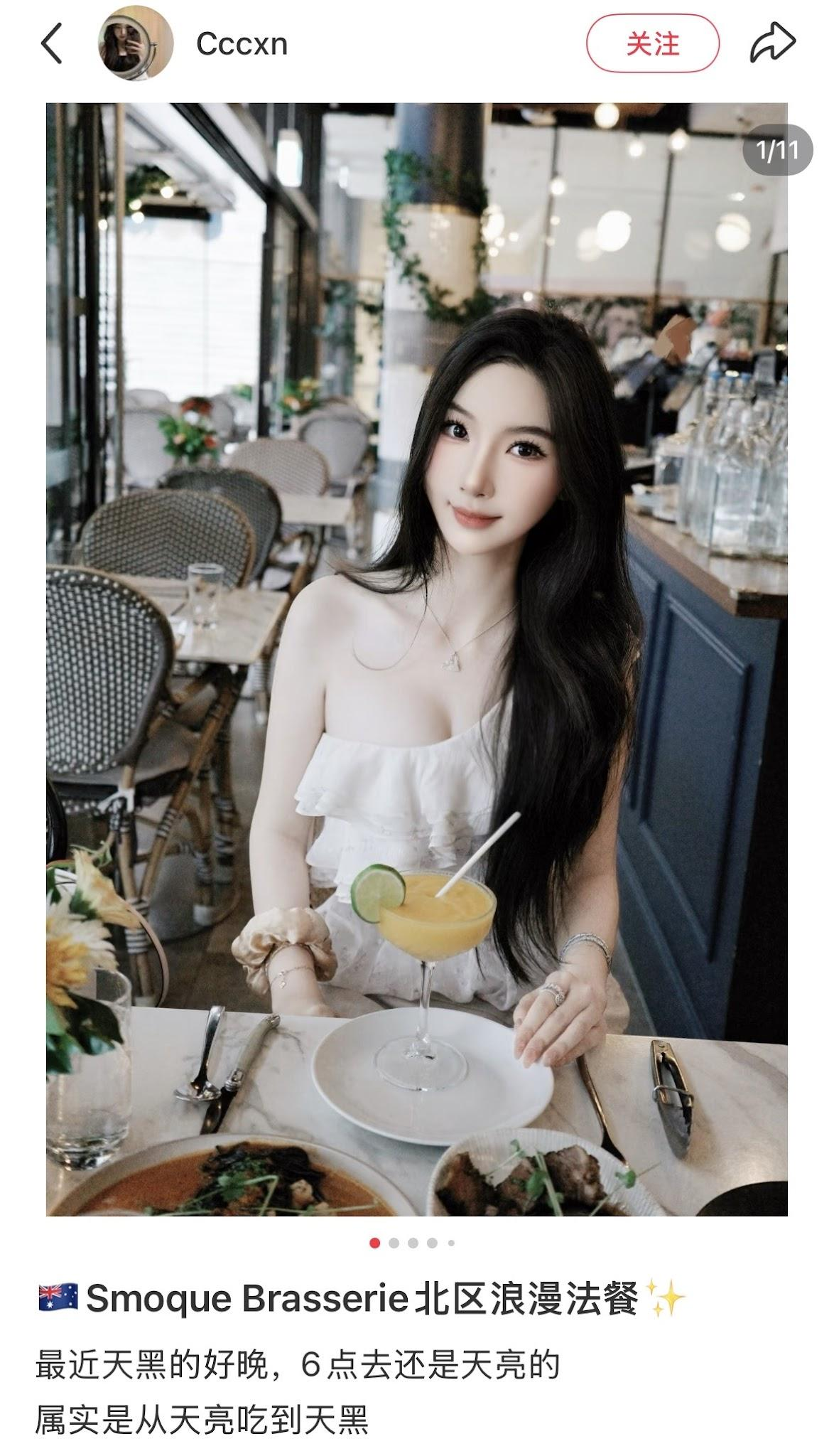 Young woman with long black hair and light skin sitting at a restaurant table with a cocktail, surrounded by plates of food. The restaurant has a modern decor with chairs, flowers, and hanging lights.