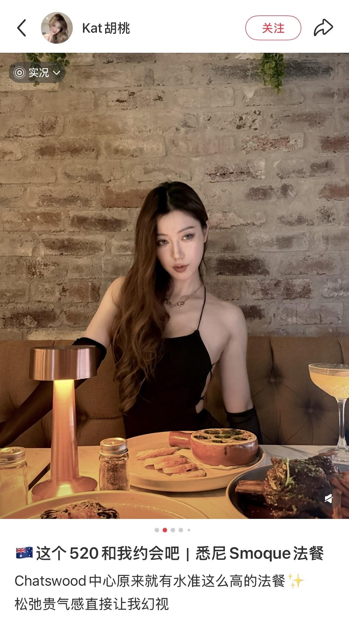 A woman with long, wavy hair wearing a black dress with cutouts, sitting at a restaurant table with food and drinks, against a exposed brick wall.