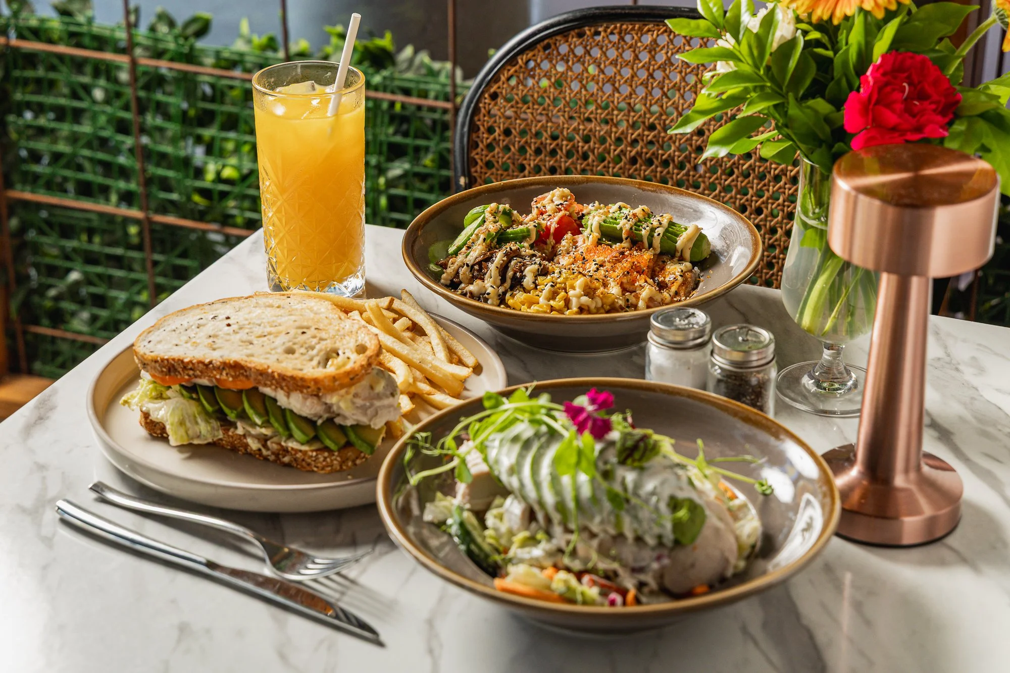 A table set with a salad, a sandwich with lettuce and tomato, a glass of orange juice, and small containers of condiments, with a background of green foliage and a flower arrangement.