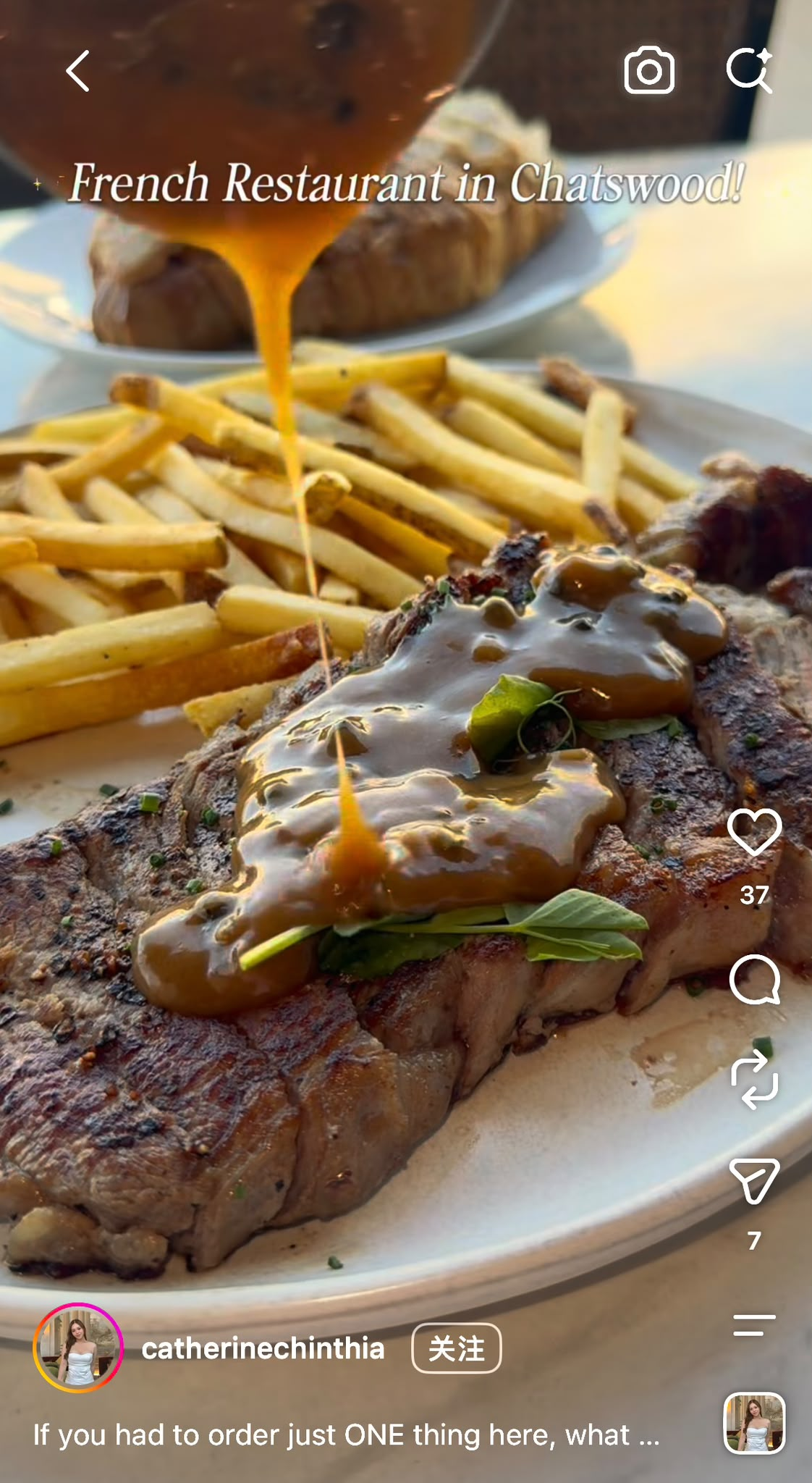 Close-up of a grilled steak topped with sauce and garnished with green herbs, served with French fries, with a glass of dark beverage in the background.