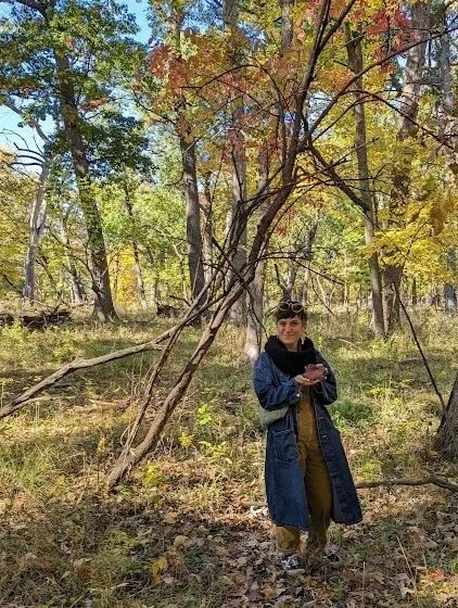 A woman standing in a forested area during autumn, surrounded by colorful fall foliage, holding a phone.