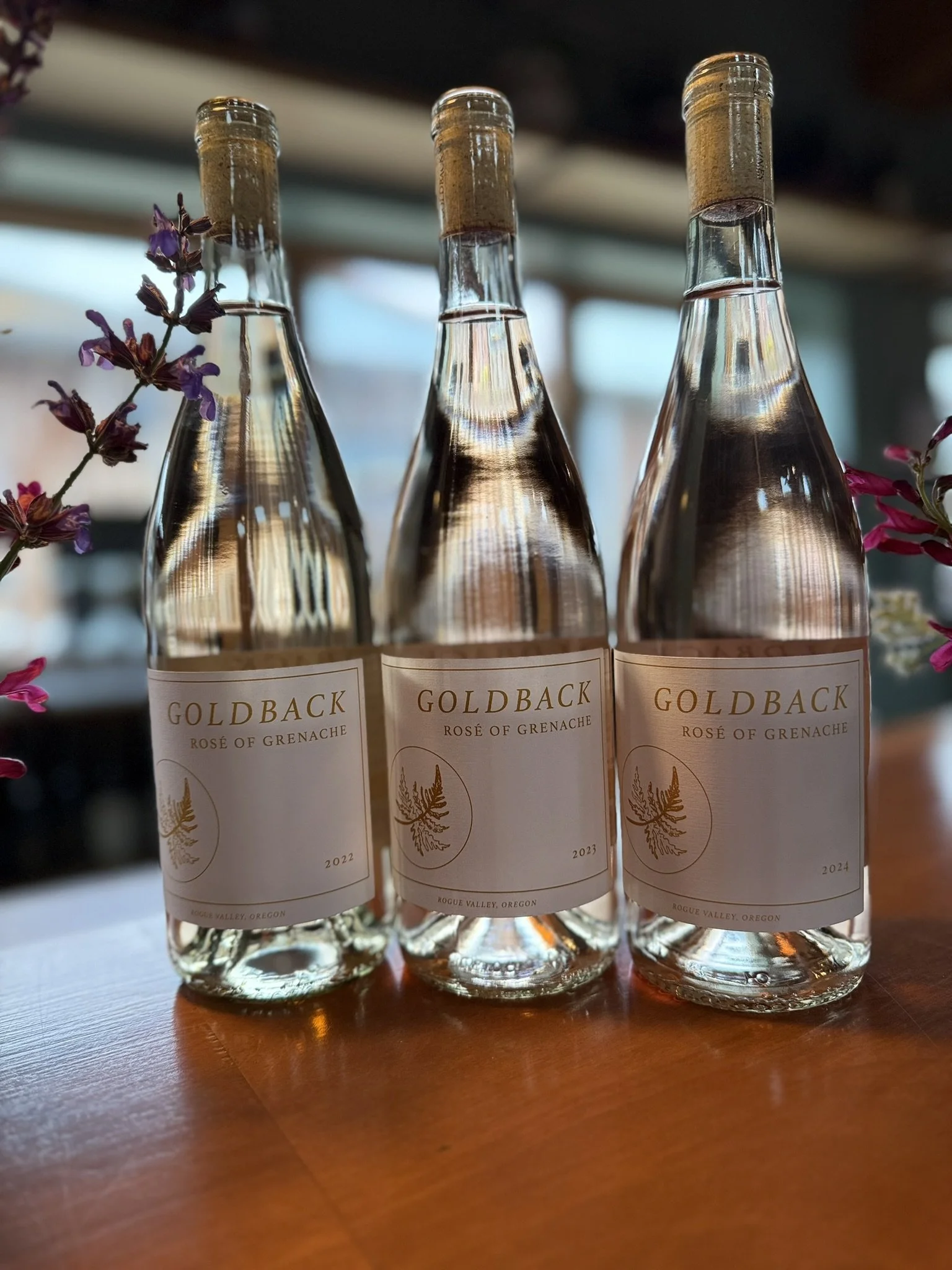 Three bottles of Goldback rosé of Grenache wine from Rogue Valley, Oregon, with vintage years 2022, 2023, and 2024, placed on a wooden surface, with some purple flowers visible on the left side.