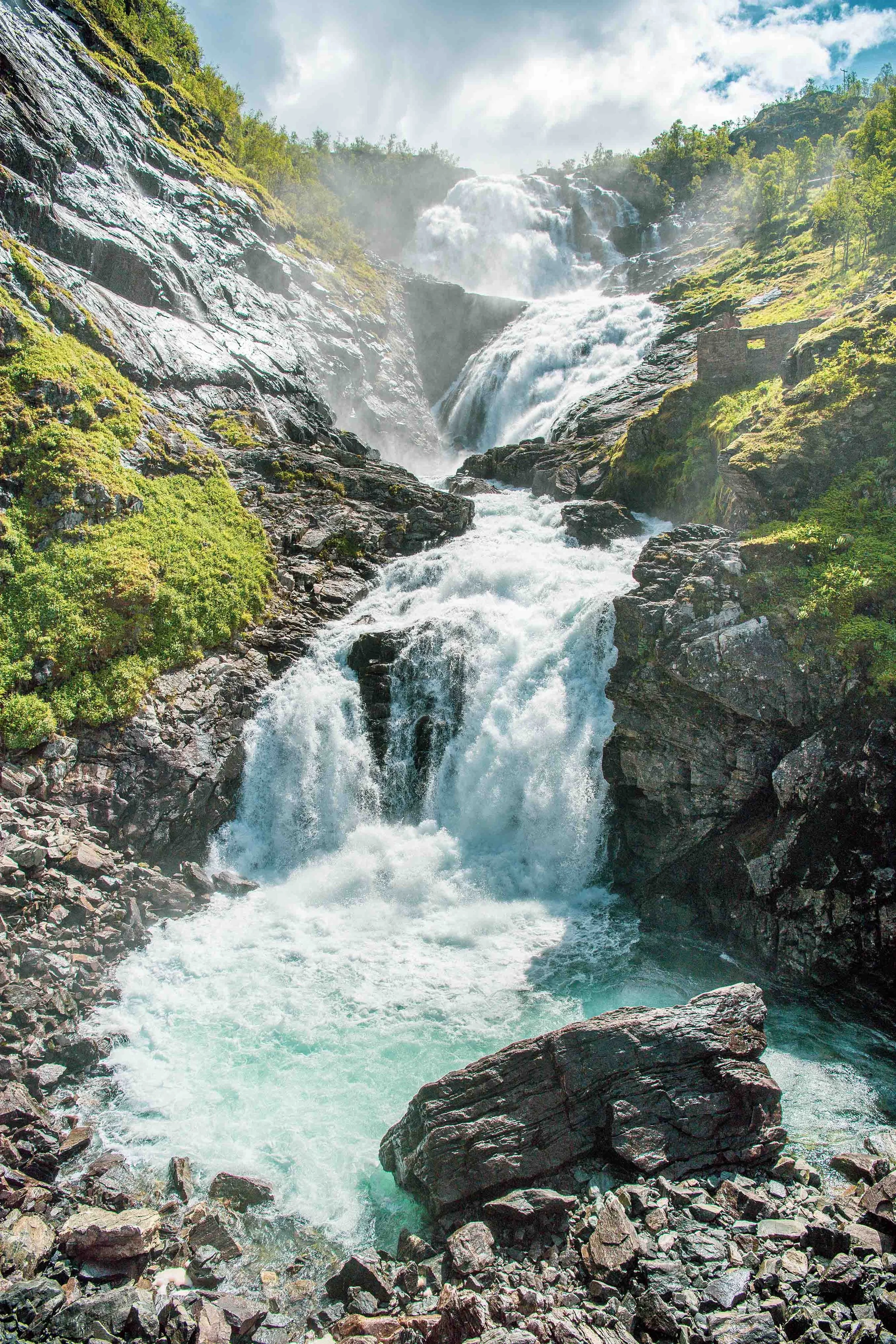 Foss med flere fossefall omringet av grønne åser og skog, med vann som fosser nedover et steinete landskap.