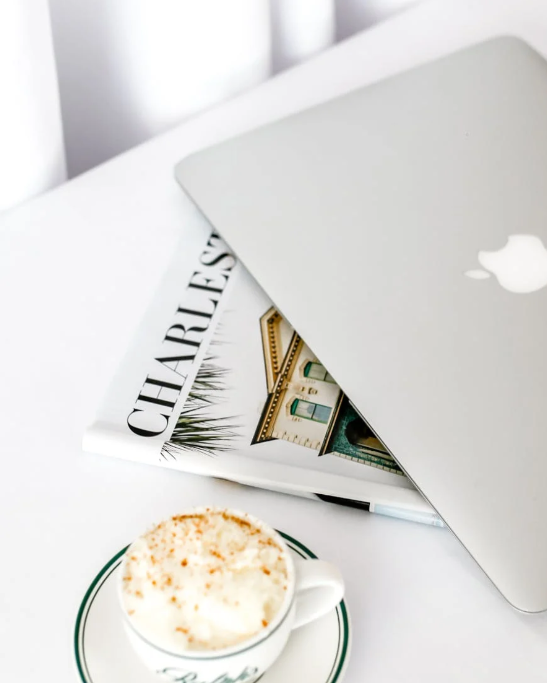 Open MacBook laptop with Apple logo, magazine titled "CHARLEST" with house images, and a cup of coffee topped with whipped cream and cinnamon on a saucer on a white table.