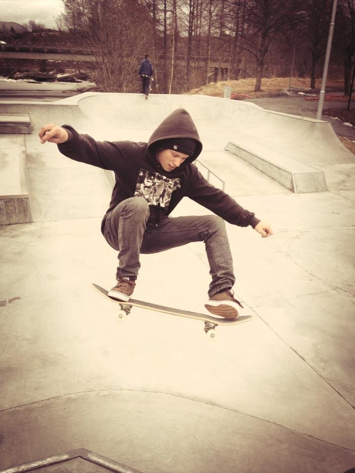 Skateur effectuant un saut dans un skatepark, portant un sweat à capuche, jeans, sneakers, en plein air.