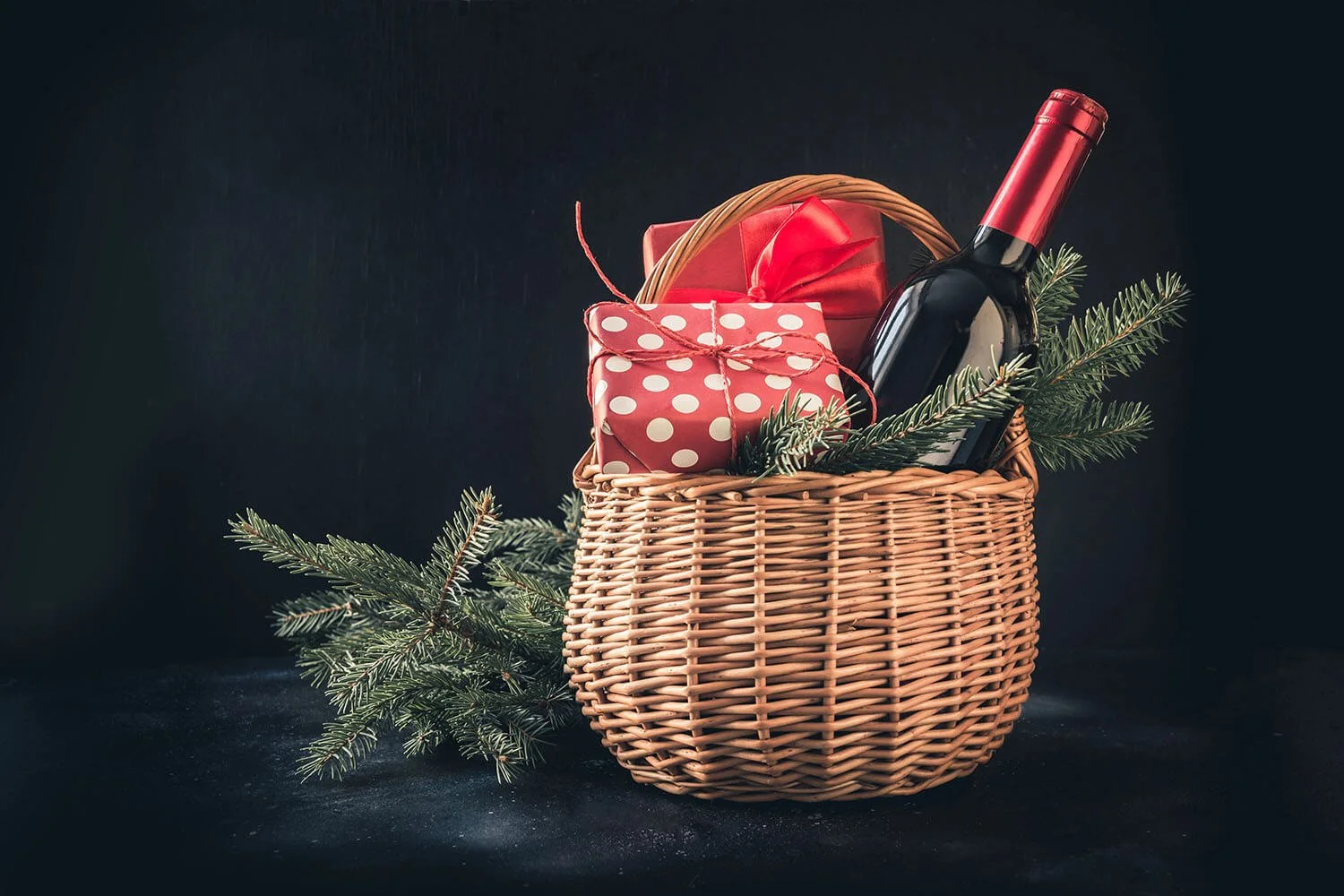 Wicker basket with wrapped gifts and a bottle of wine and pine boughs inside