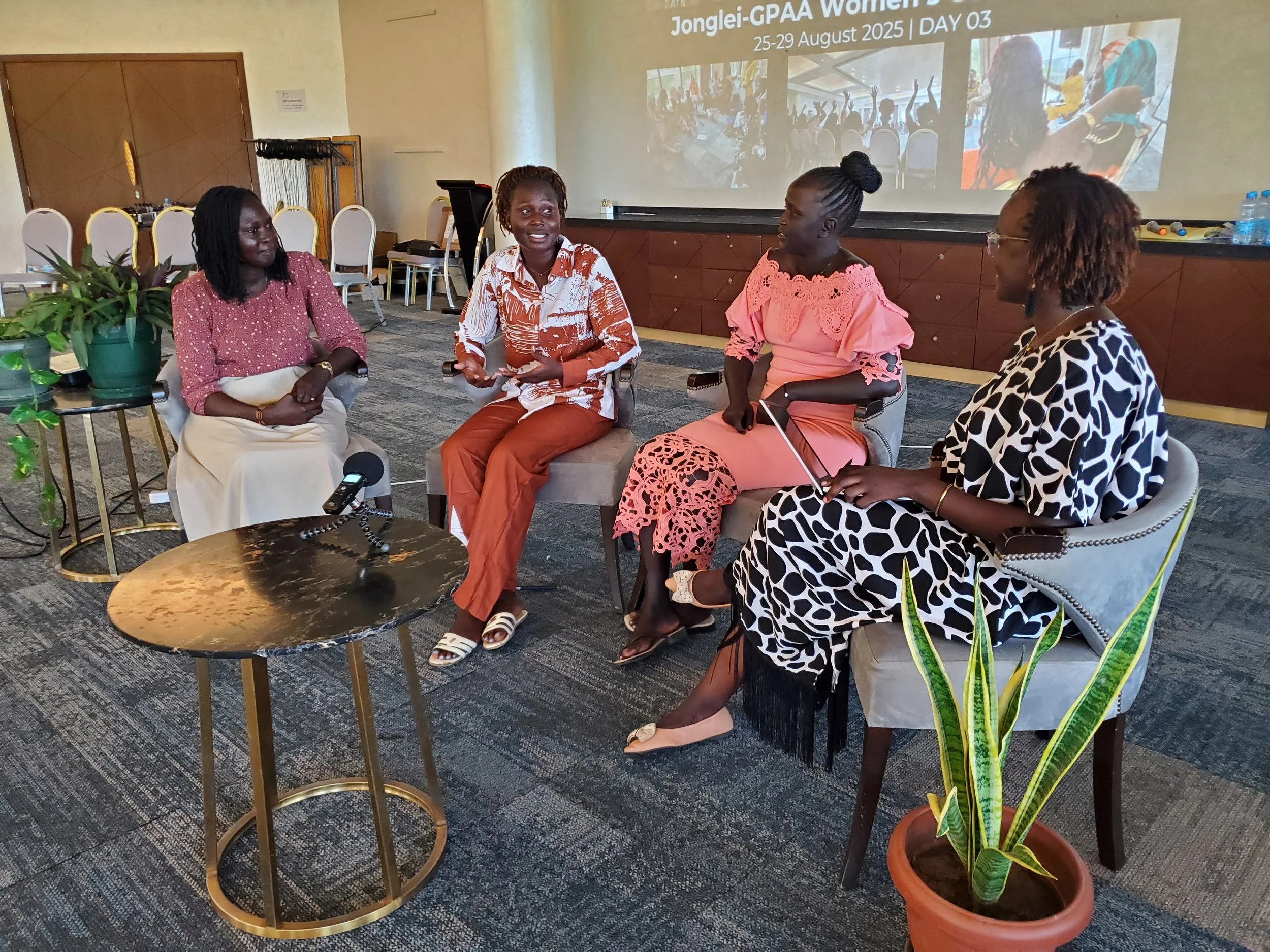 Four women seated in a panel discussion or interview setting, engaged in conversation inside a conference room, with a presentation slide in the background mentioning the Jonglei-GPAA Women event from August 25-29, 2025.