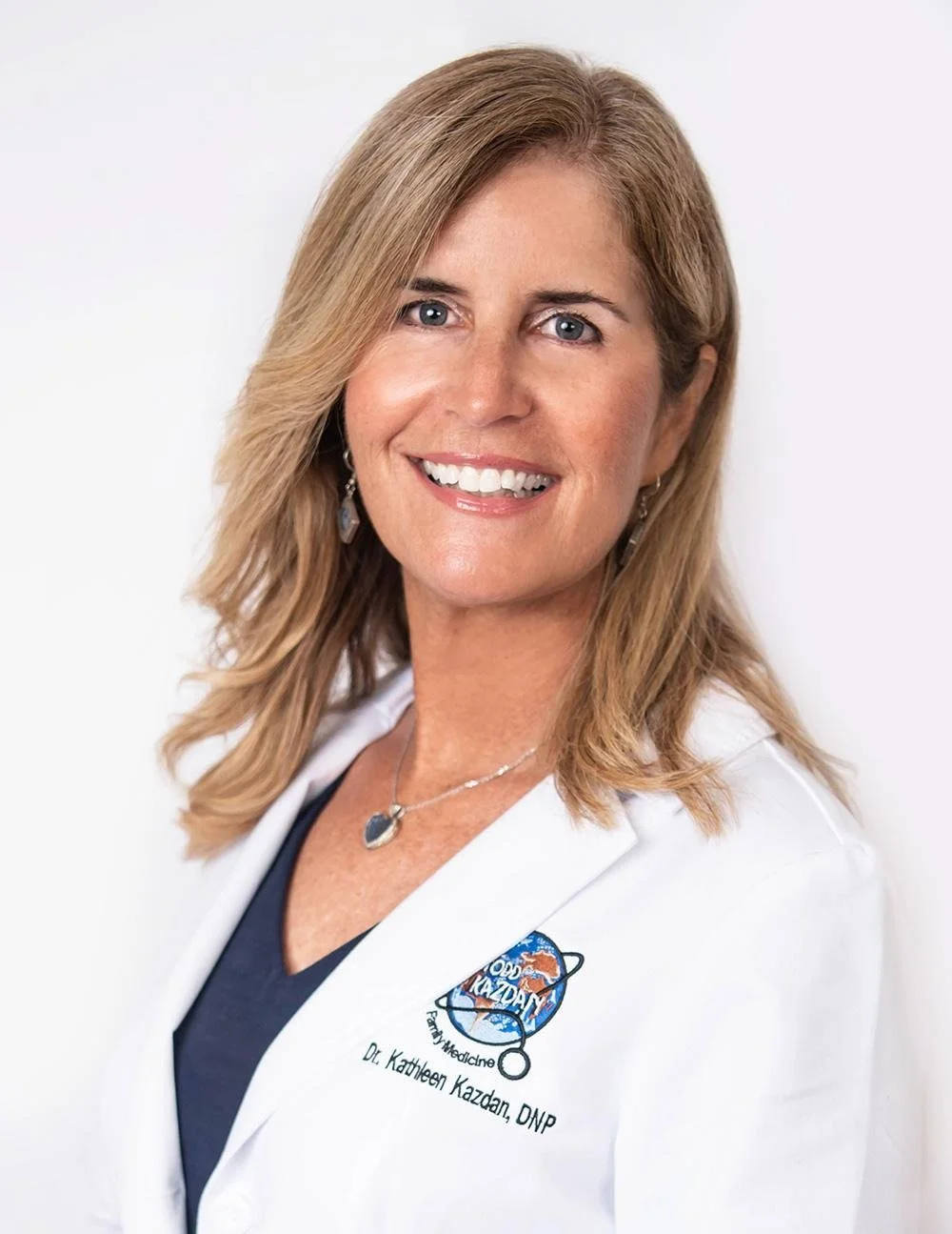A woman with blonde hair smiling, wearing a white coat with a logo, a dark top, and jewelry, against a plain white background.