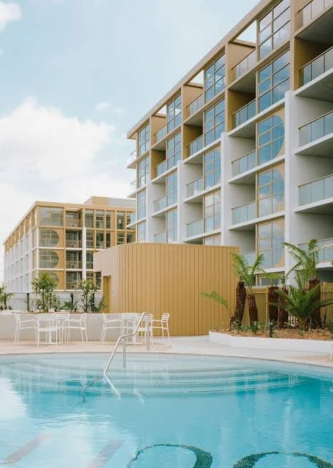 Modern apartment buildings with balconies and large windows, pool area with white lounge chairs, and palm trees in the courtyard.