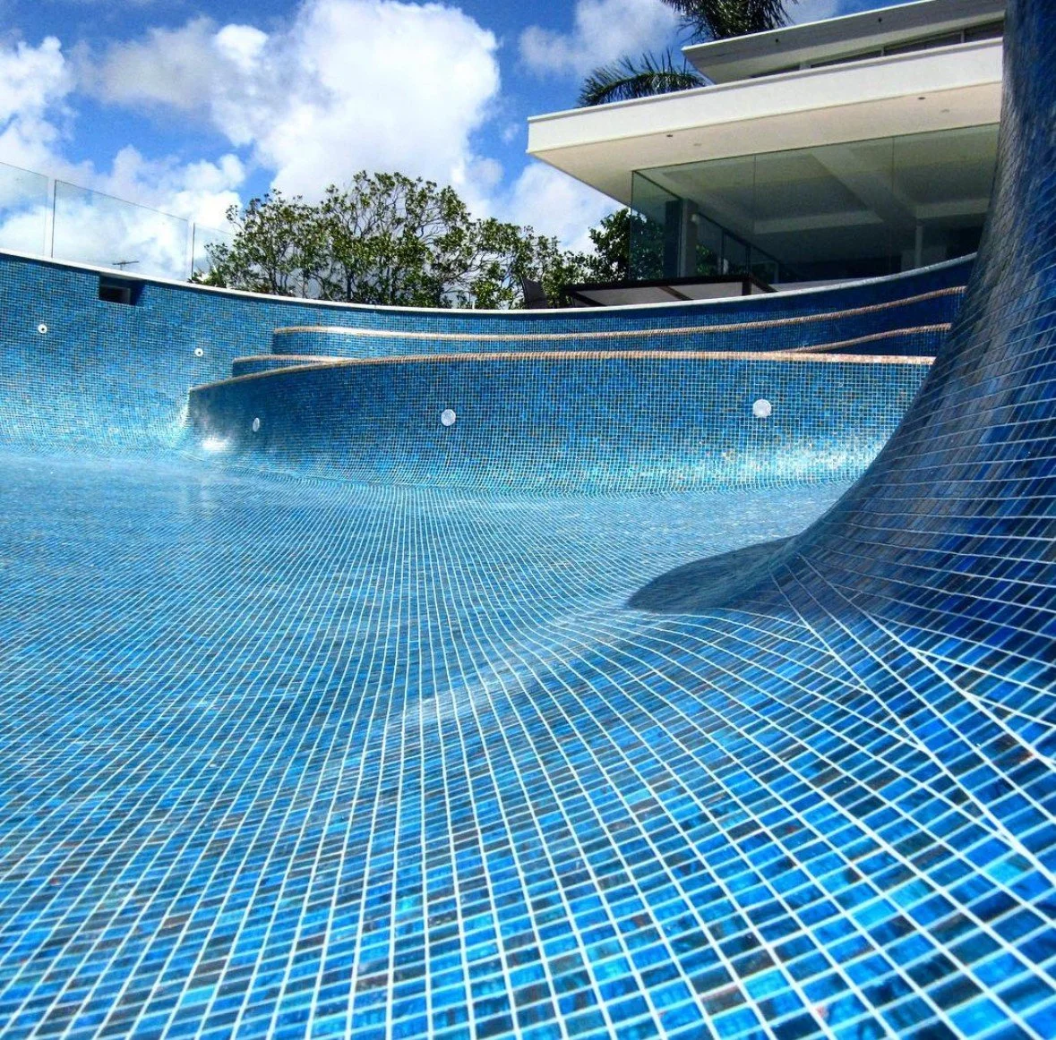 Empty outdoor swimming pool with blue mosaic tiles, steps leading into the water, and a modern building with a flat roof in the background, under a partly cloudy sky.