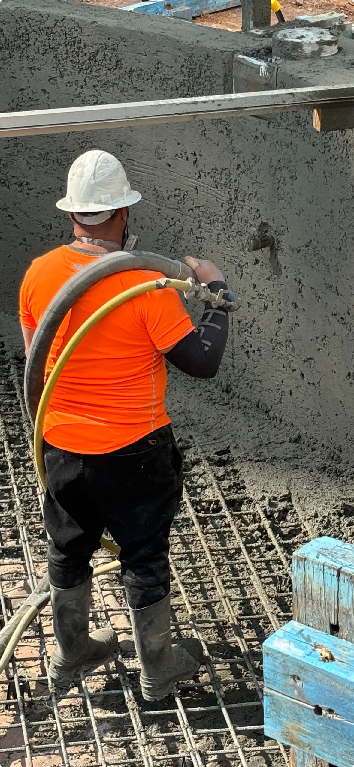 Worker wearing an orange shirt, a hard hat, and boots, applying or smoothing wet concrete on a construction site, with visible rebar and a blue wooden block nearby.