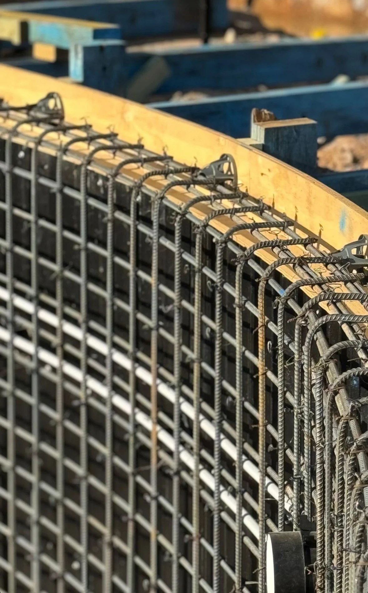 Close-up of a steel rebar grid with a yellow wooden form and blue wooden framing in the background, at a construction site.