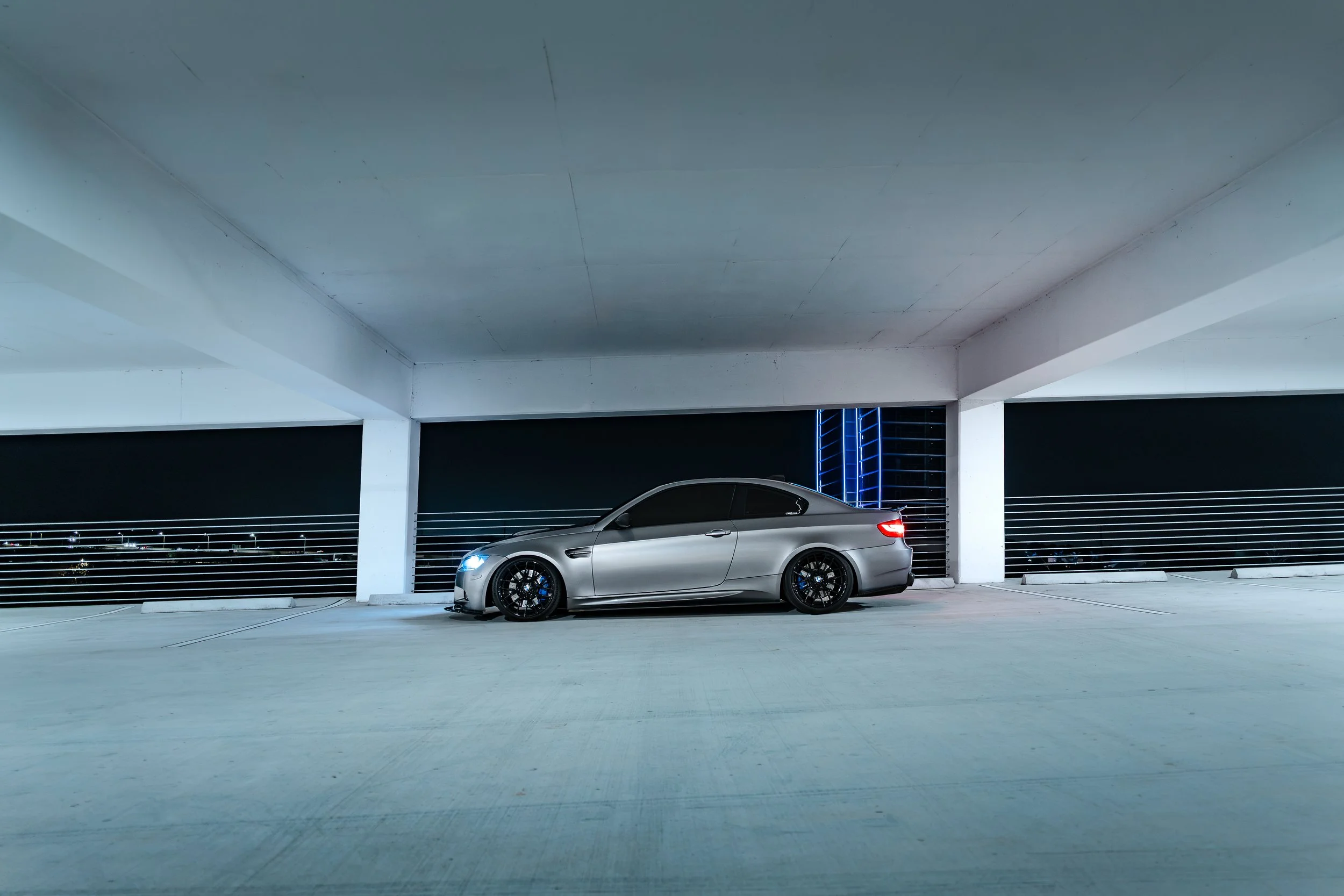 A silver sports car with tinted windows parked in an empty, multi-level parking garage at night, illuminated by nearby city lights and blue neon accents.