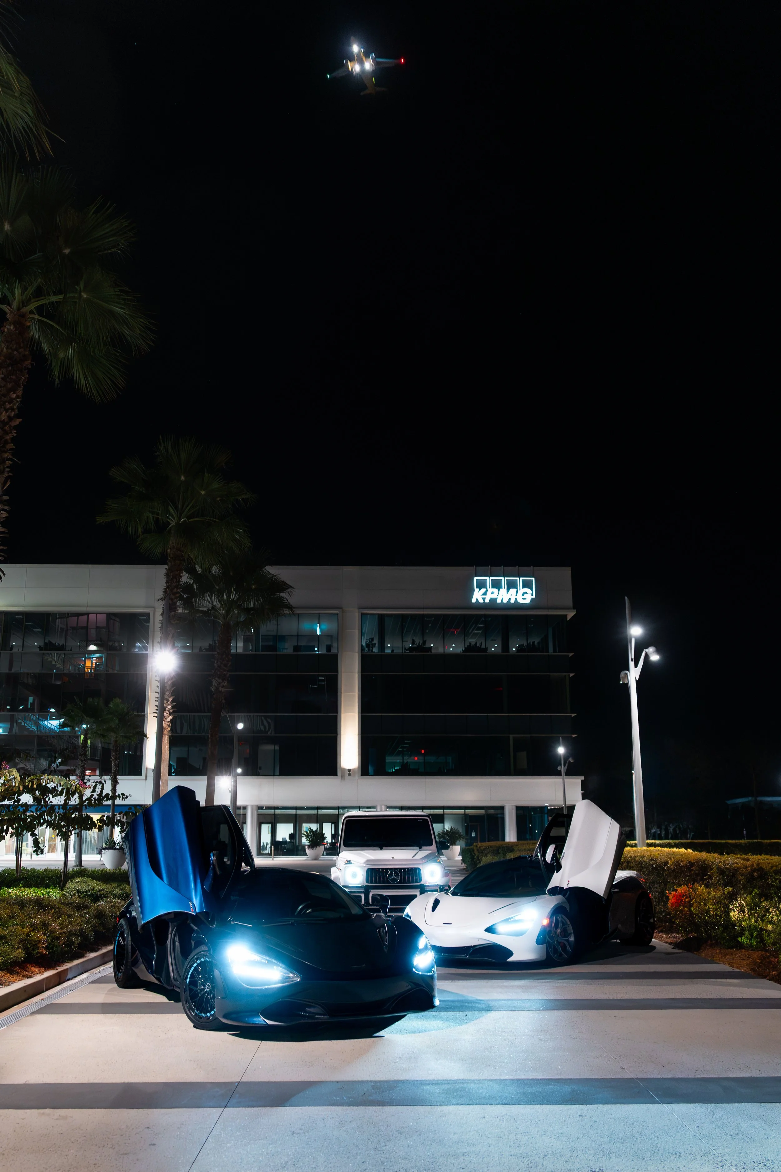 Nighttime scene featuring two luxury sports cars with vertically opening doors, a white SUV, and a modern office building with a KPMG logo, palm trees, and a helicopter flying above.