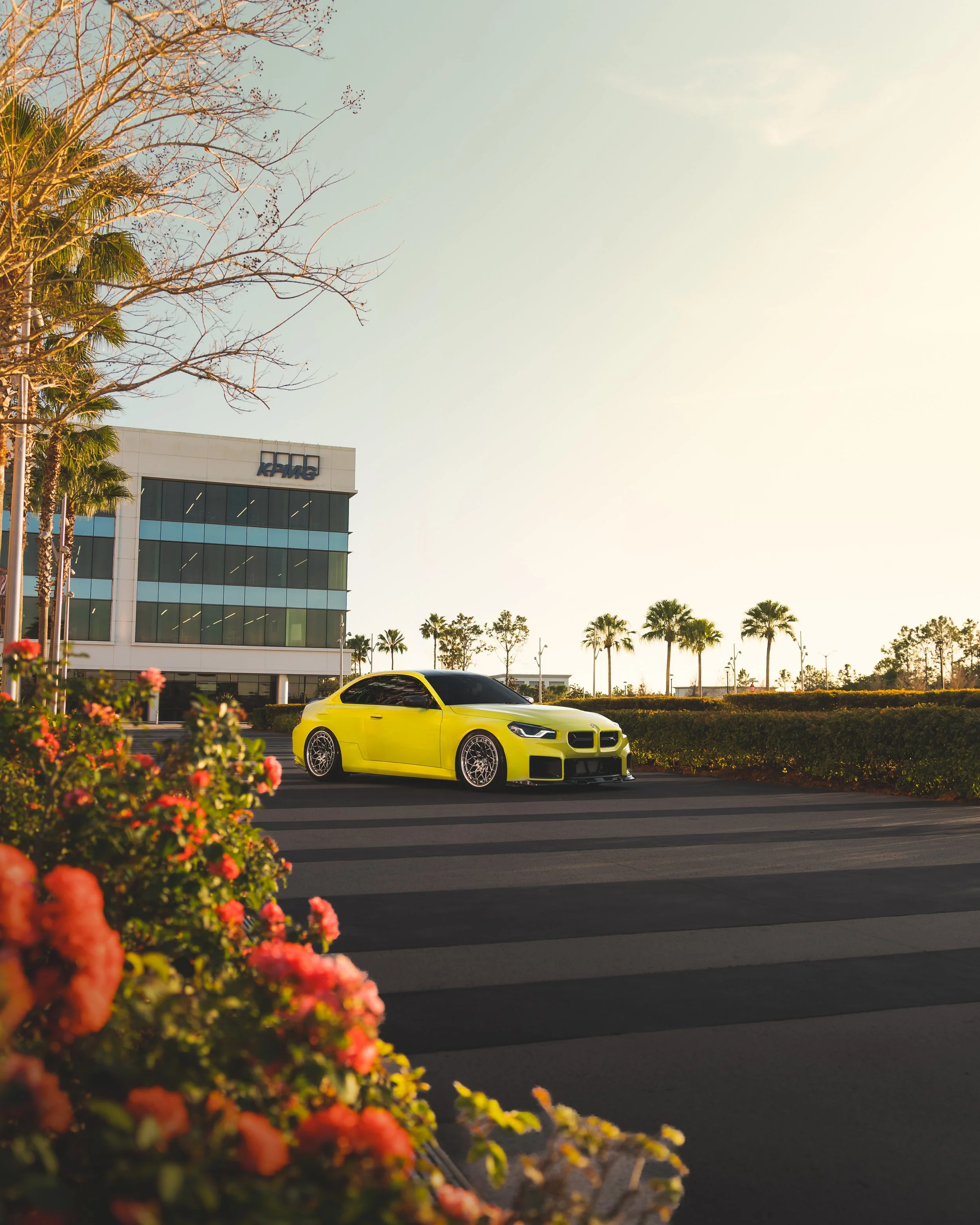 A bright yellow sports car parked on a street near a modern office building with the sign 'KPMG', with palm trees and bushes in the background, during sunset or sunrise.