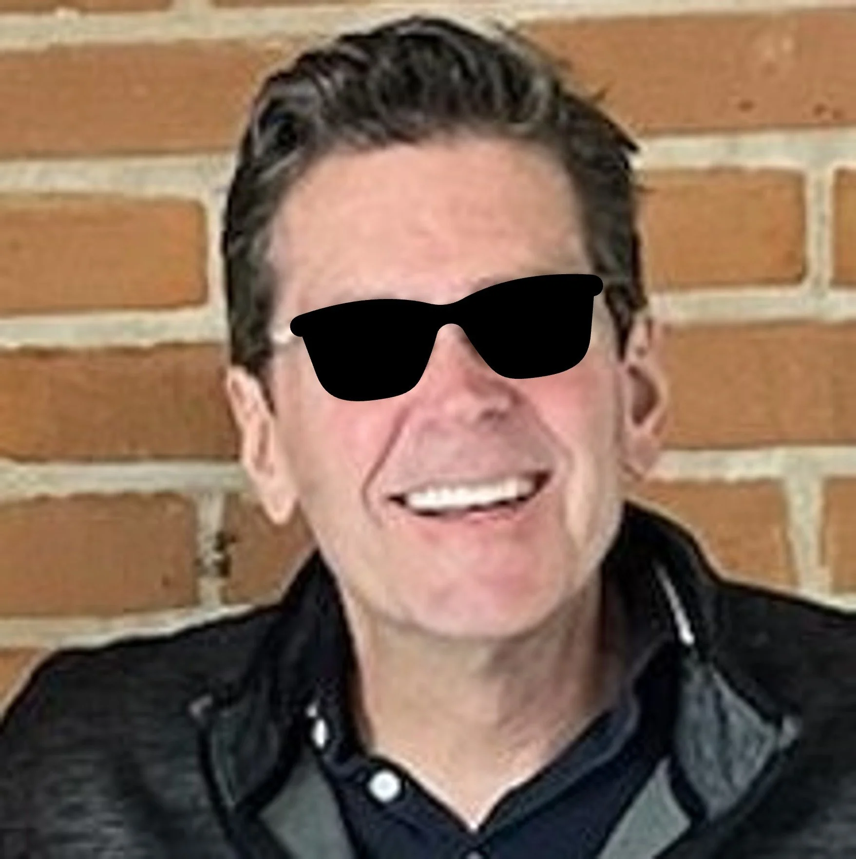 A person with dark, styled hair wearing sunglasses, a black jacket, and a gray shirt, smiling in front of a brick wall.