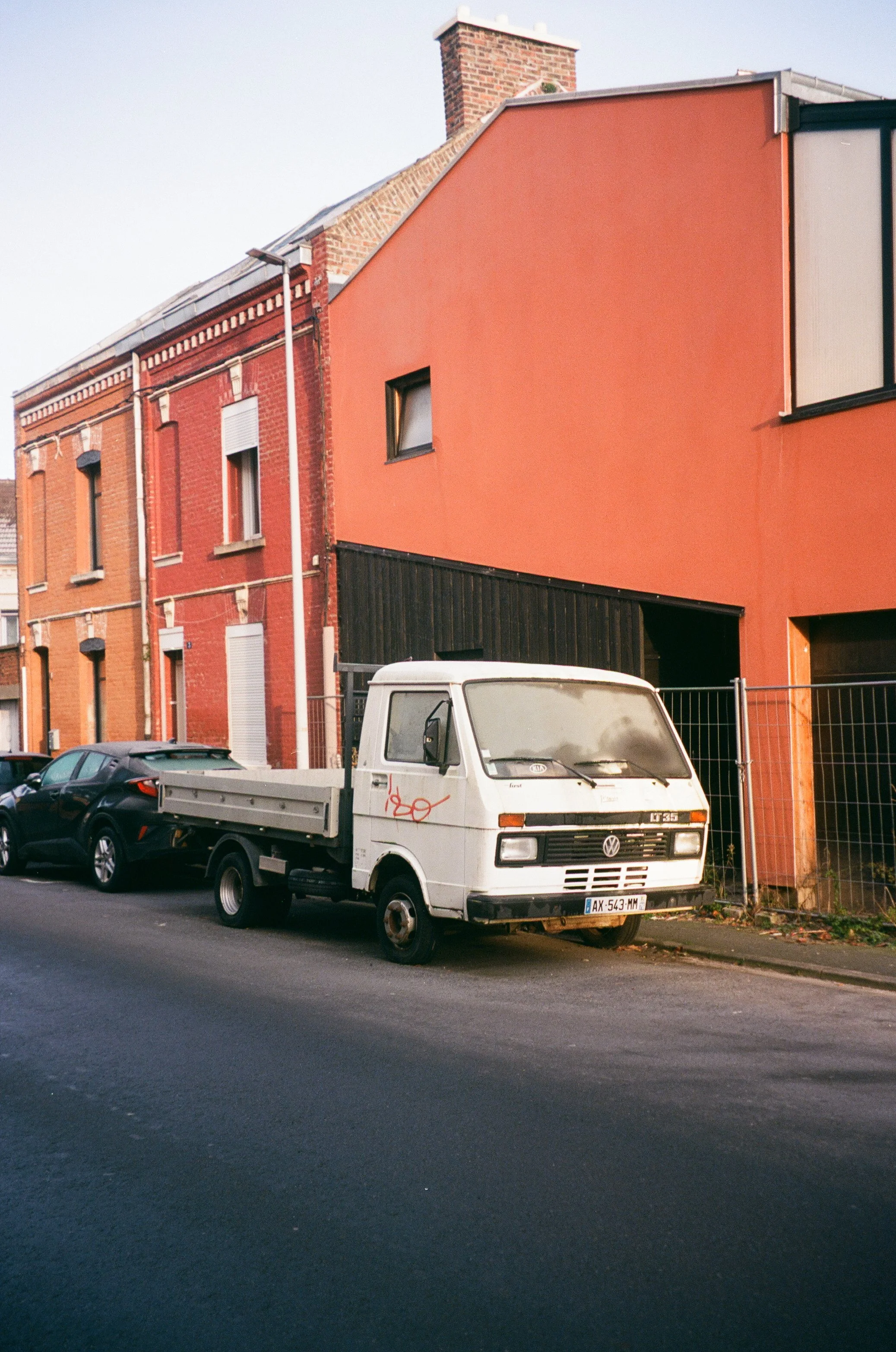 Older white Volkswagen LT35 truck parked on the street, in front of a modern pink and black building and a row of red brick houses.