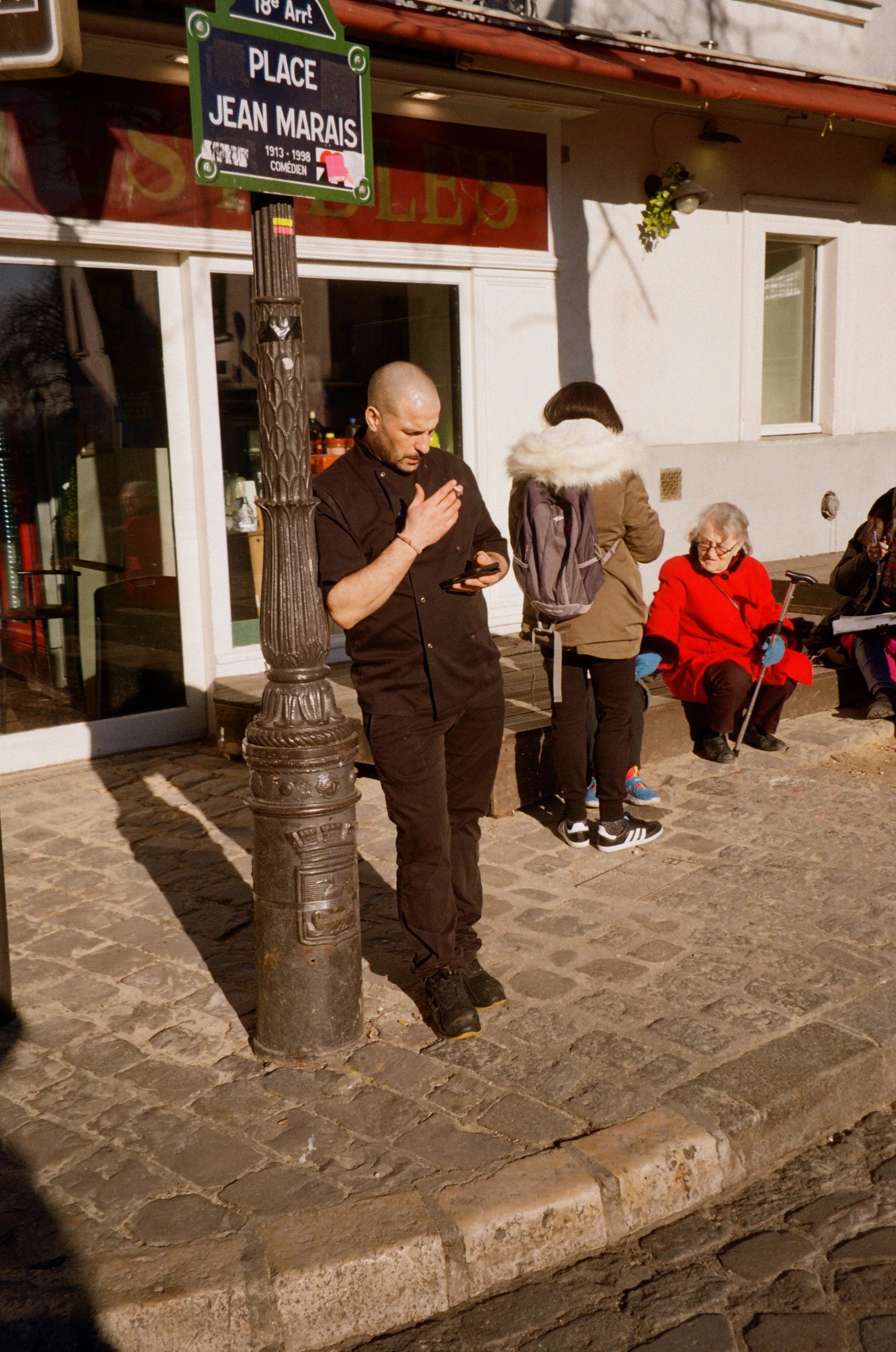 A man in black clothes standing on cobblestone sidewalk, looking at his phone, next to a streetlamp, with people seated on a bench nearby.