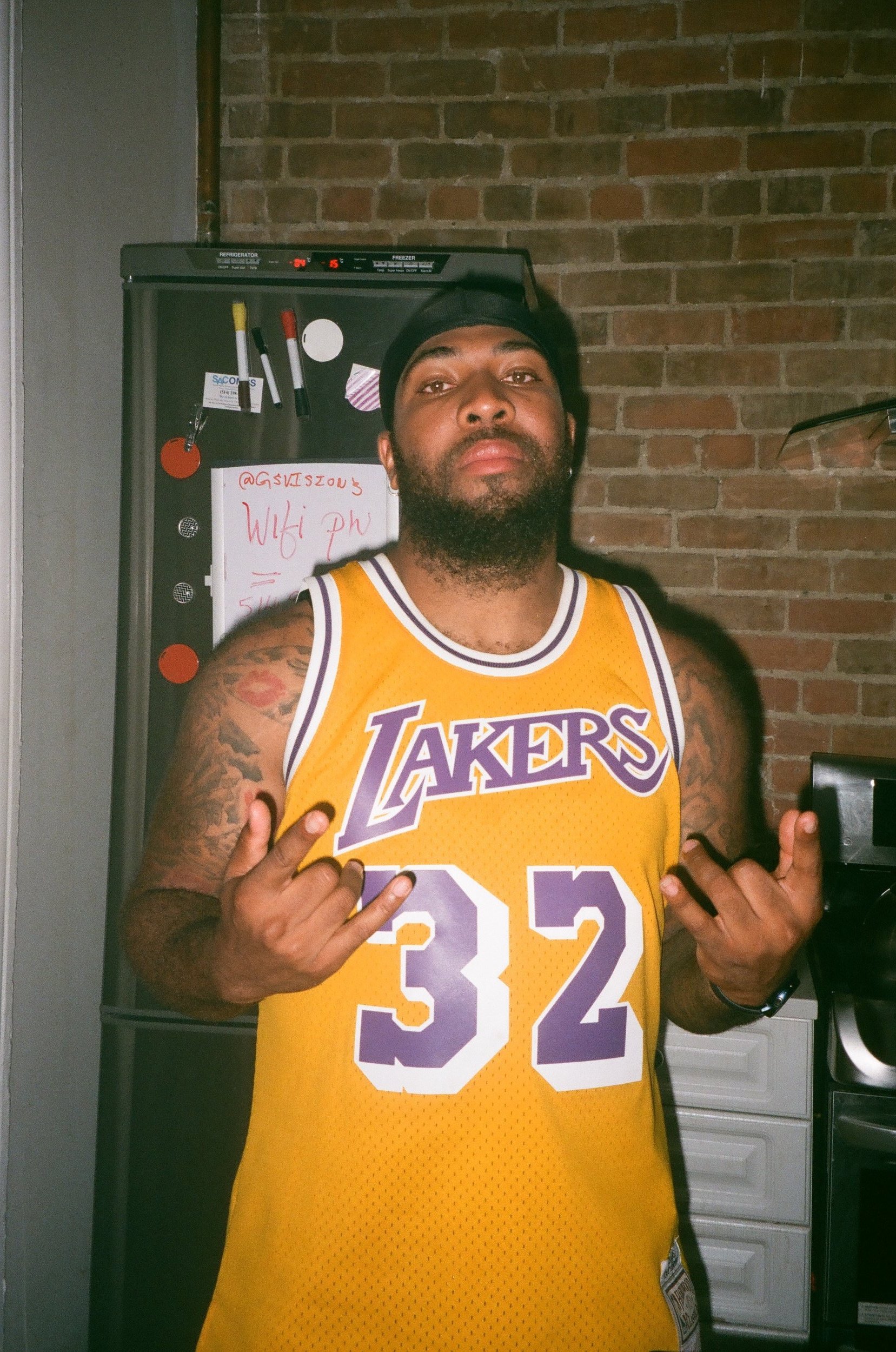 Man wearing a yellow Los Angeles Lakers basketball jersey with the number 32, making a hand gesture. Standing in front of a refrigerator and a brick wall.