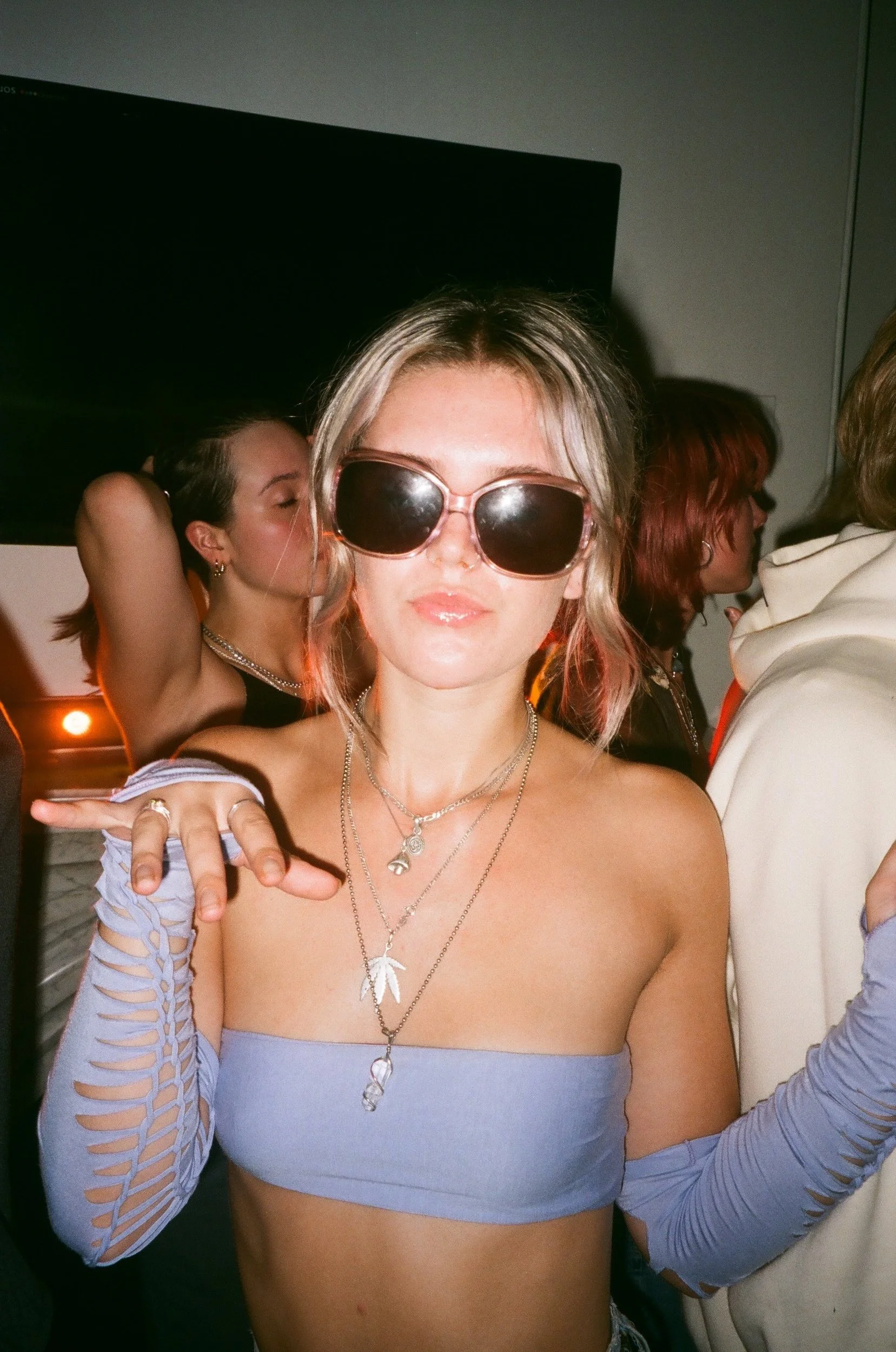 A young woman with blonde hair wearing large sunglasses, layered necklaces, and a light blue off-shoulder top with long, cut-out sleeves, at a party.