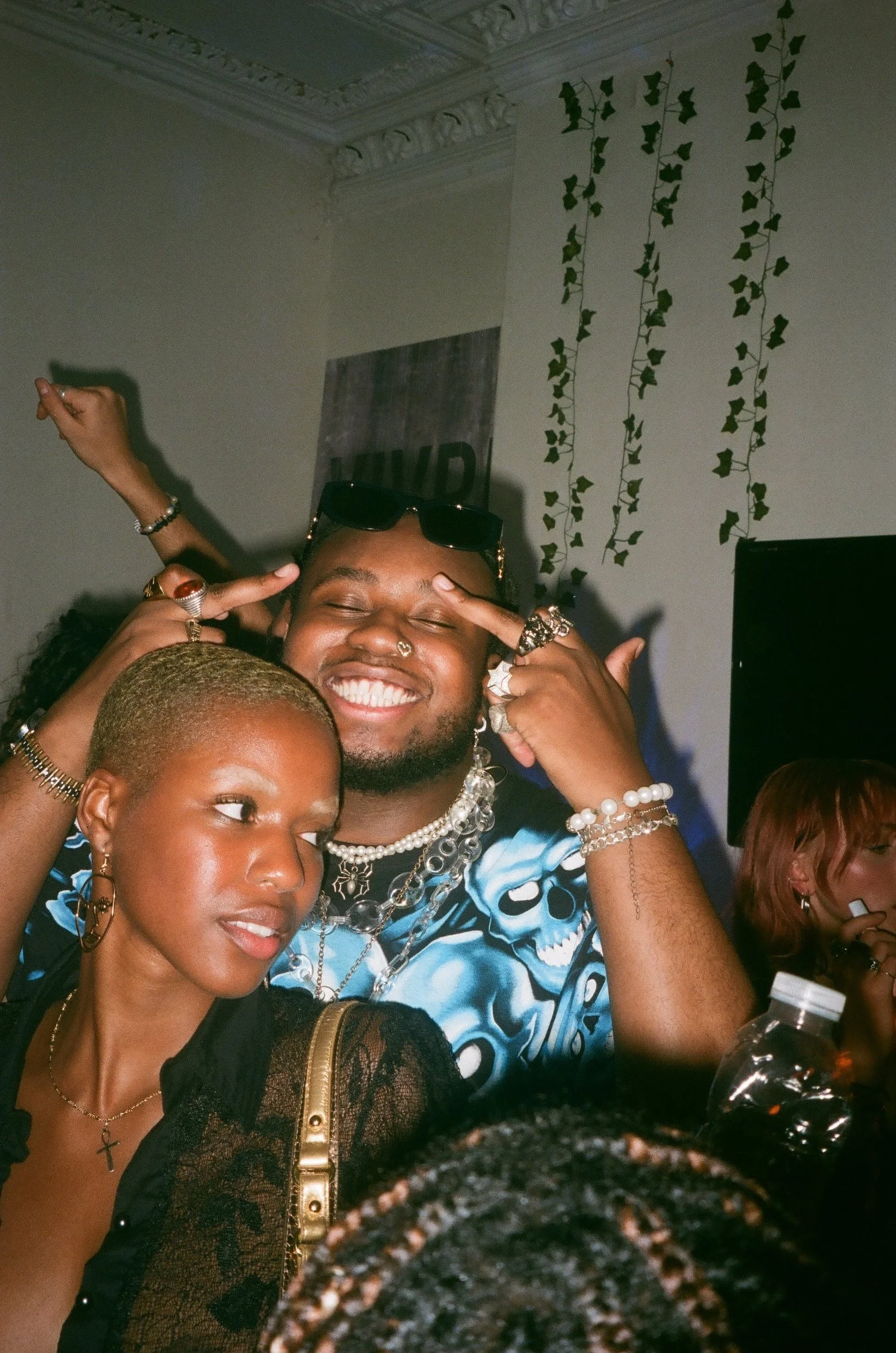 A group of people at a party, with two individuals in the forefront smiling and posing for the camera. The person on the left has short blonde hair and wears jewelry, while the person on the right has dark hair, sunglasses on their head, and is weari