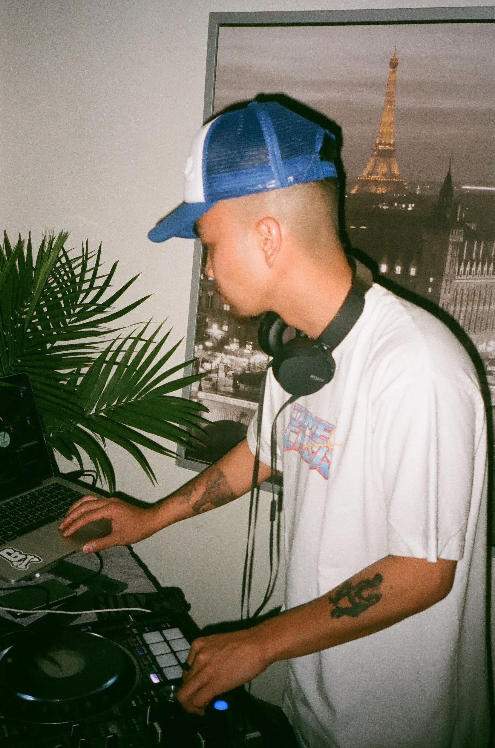 A young man wearing a white t-shirt with a graphic, a blue and white trucker hat, and headphones around his neck is DJing with a laptop and DJ controller in front of him. There is a poster of Paris with Eiffel Tower in the background and a green plan