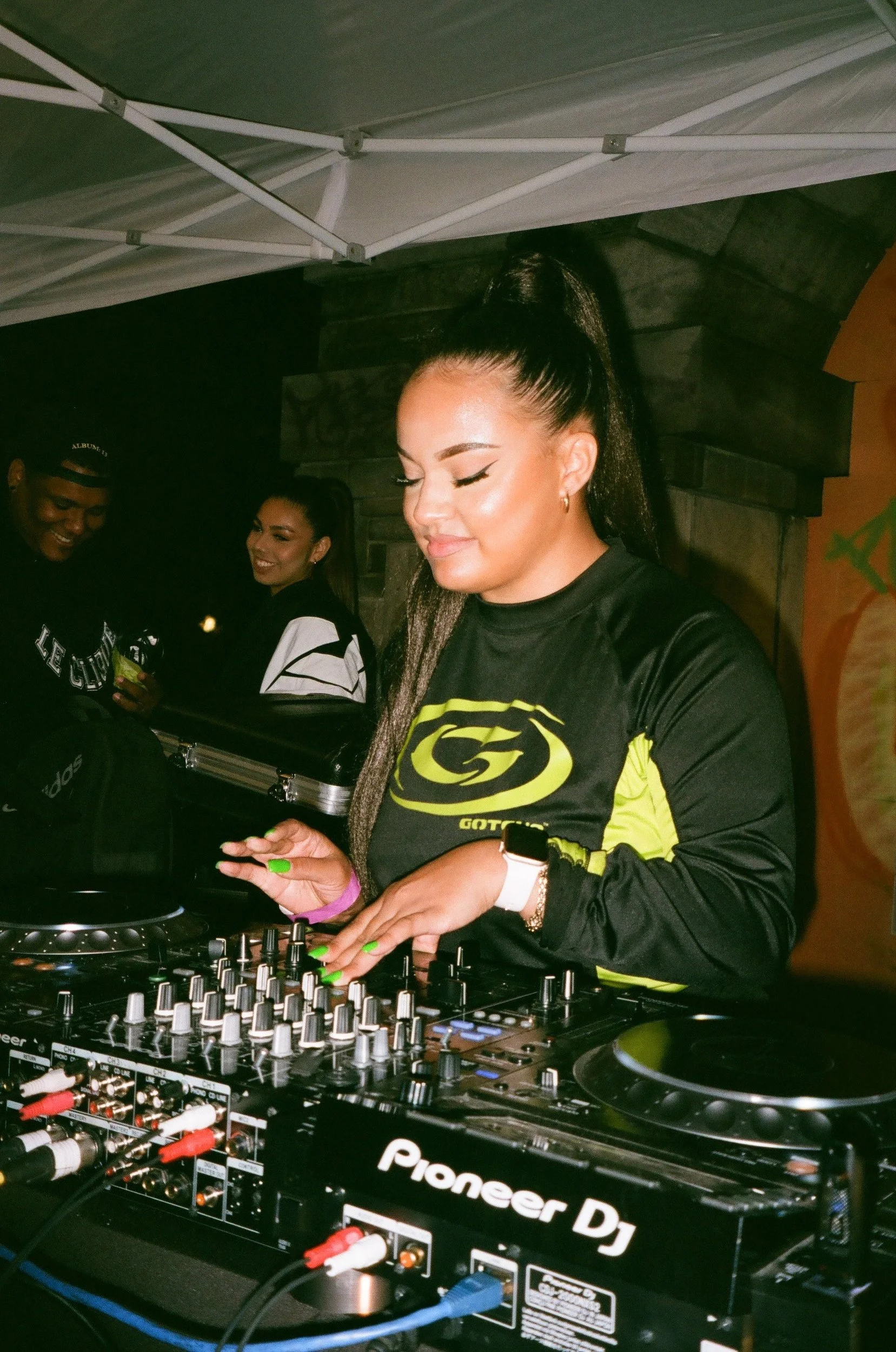 A woman DJ with long dark hair in a ponytail, wearing a black and yellow athletic shirt, working at a Pioneer DJ controller amidst other people at a party or club setting.