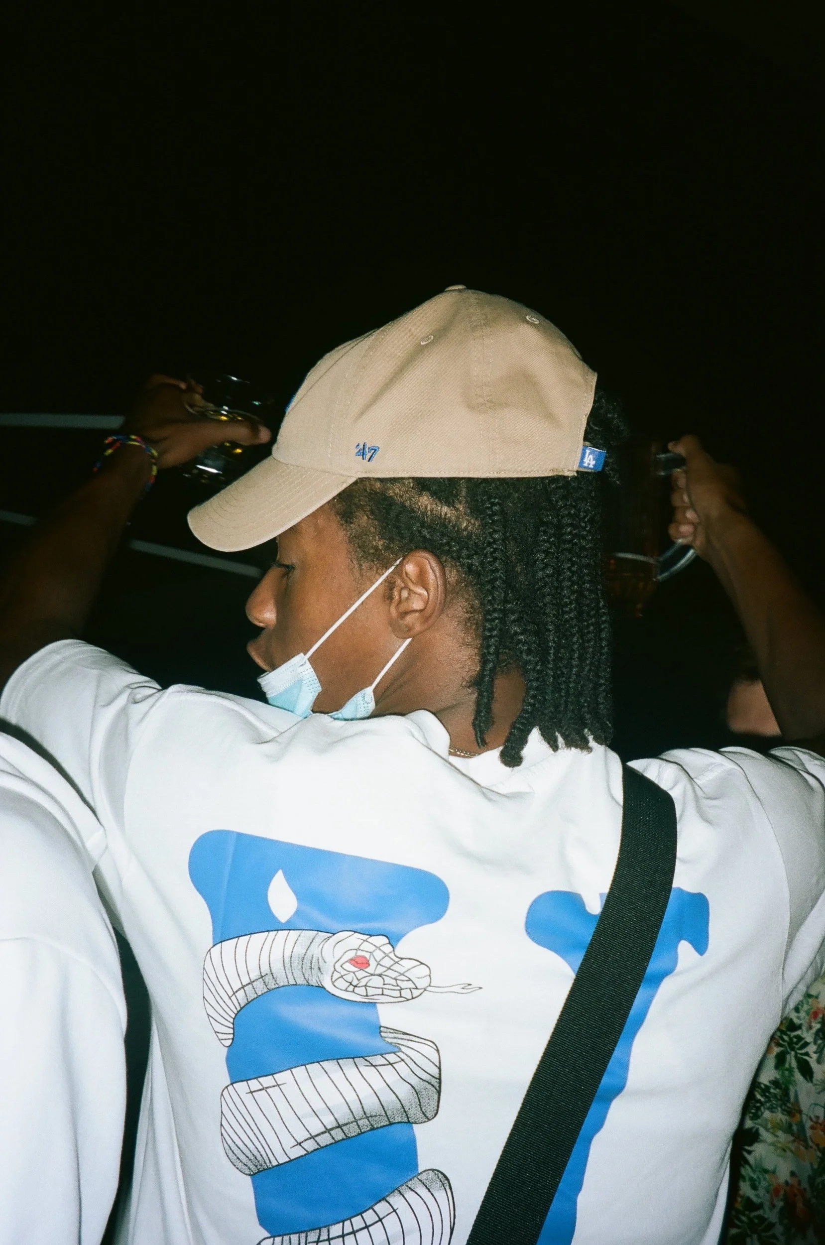 Person with braided hair wearing a beige cap and a white T-shirt with a snake graphic, holding a drink, at night.