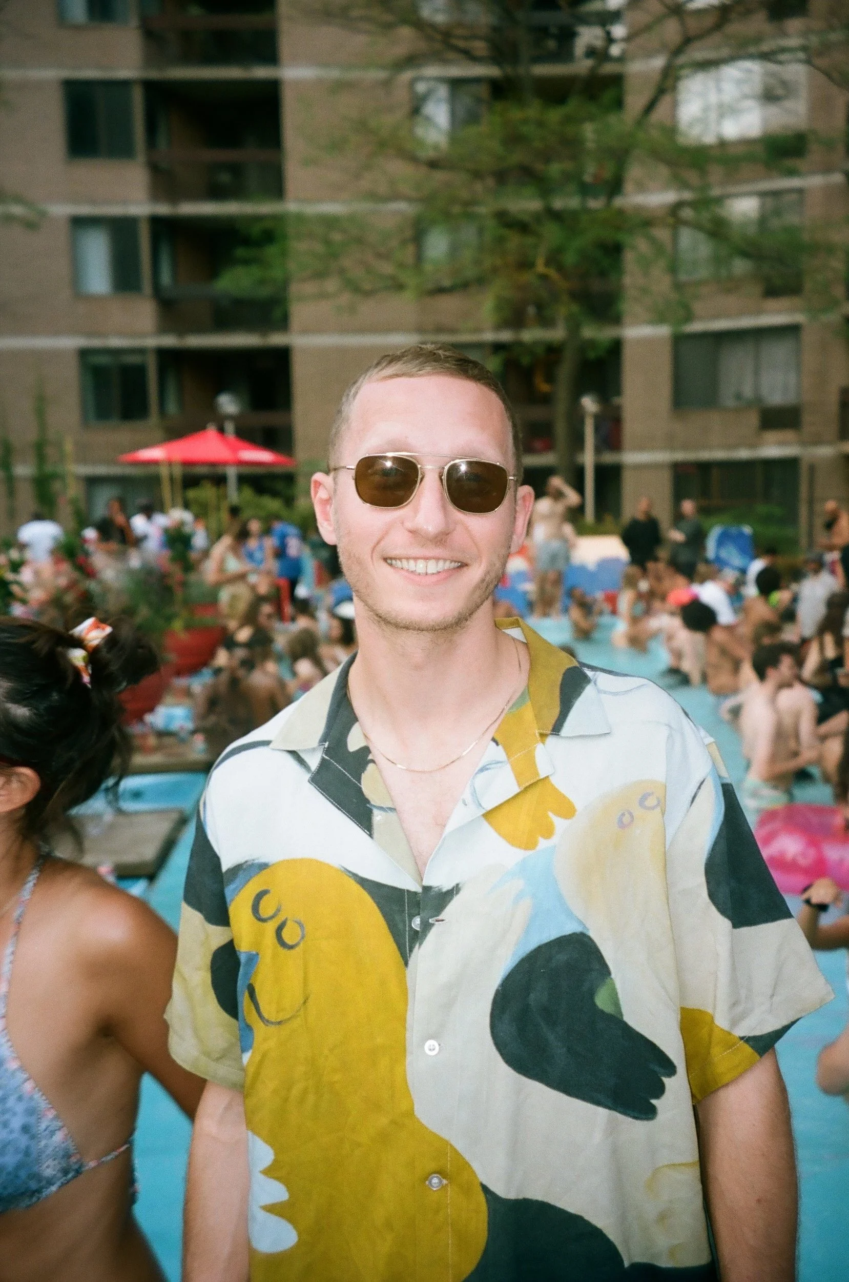 A smiling man wearing sunglasses and a colorful Hawaiian shirt at a pool party, with many people in swimsuits in the background.