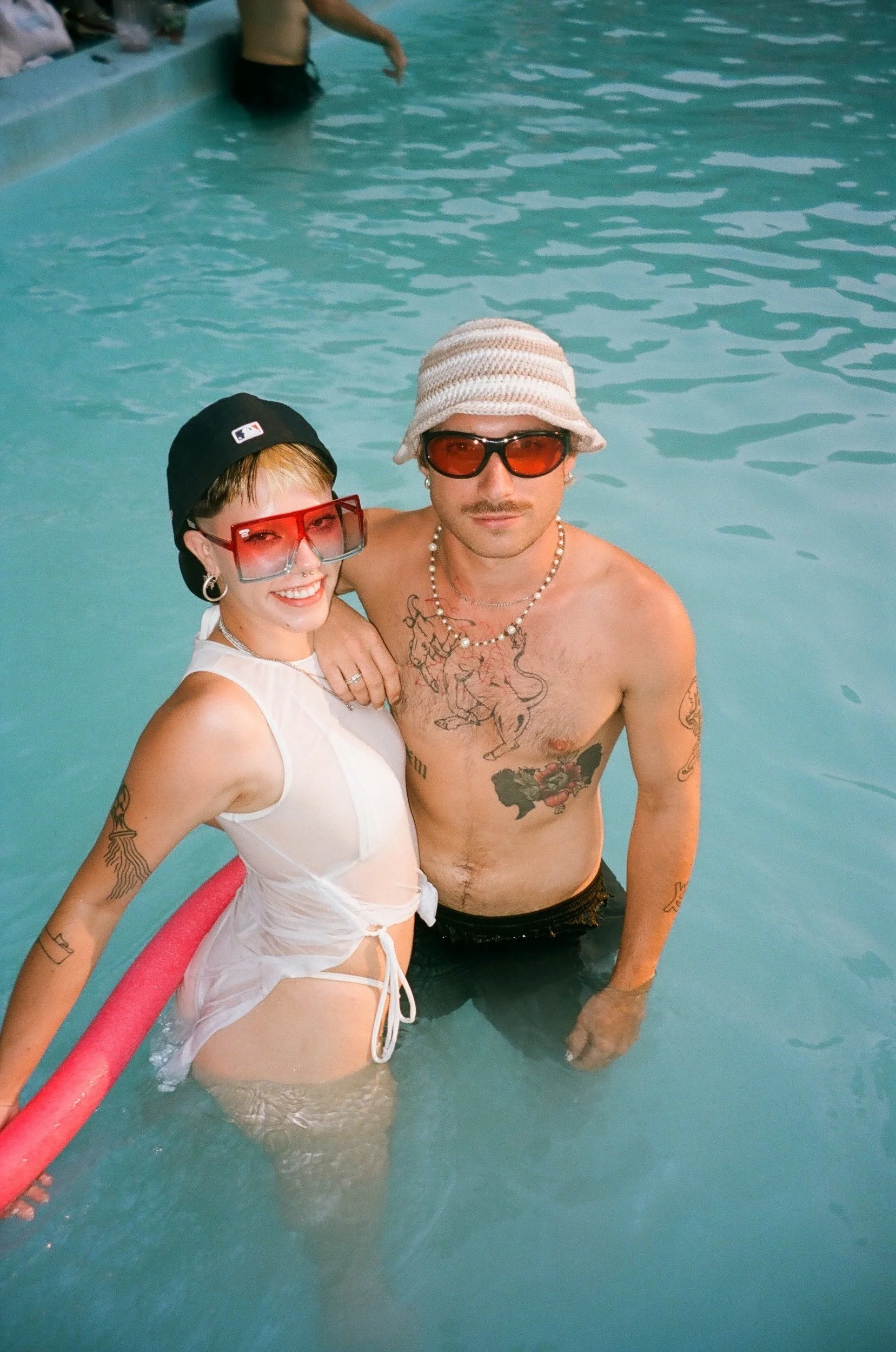 Two people standing in a swimming pool, wearing sunglasses and hats, smiling at the camera. The woman has a short hairstyle, is wearing a white mesh cover-up, and the man has tattoos and is shirtless, wearing dark swimming trunks. The woman has her a