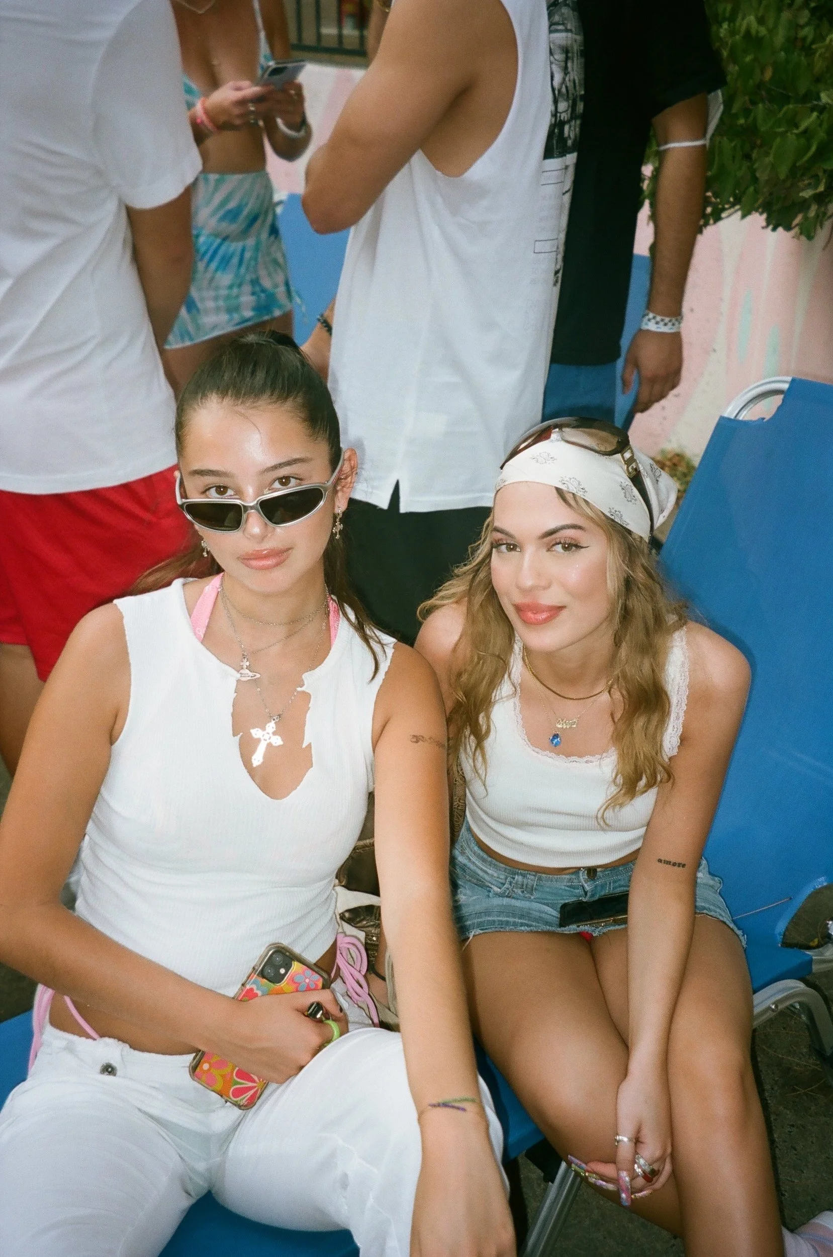 Two young women sitting outdoors, one wearing white with sunglasses and the other with a bandana, surrounded by people at a social gathering.