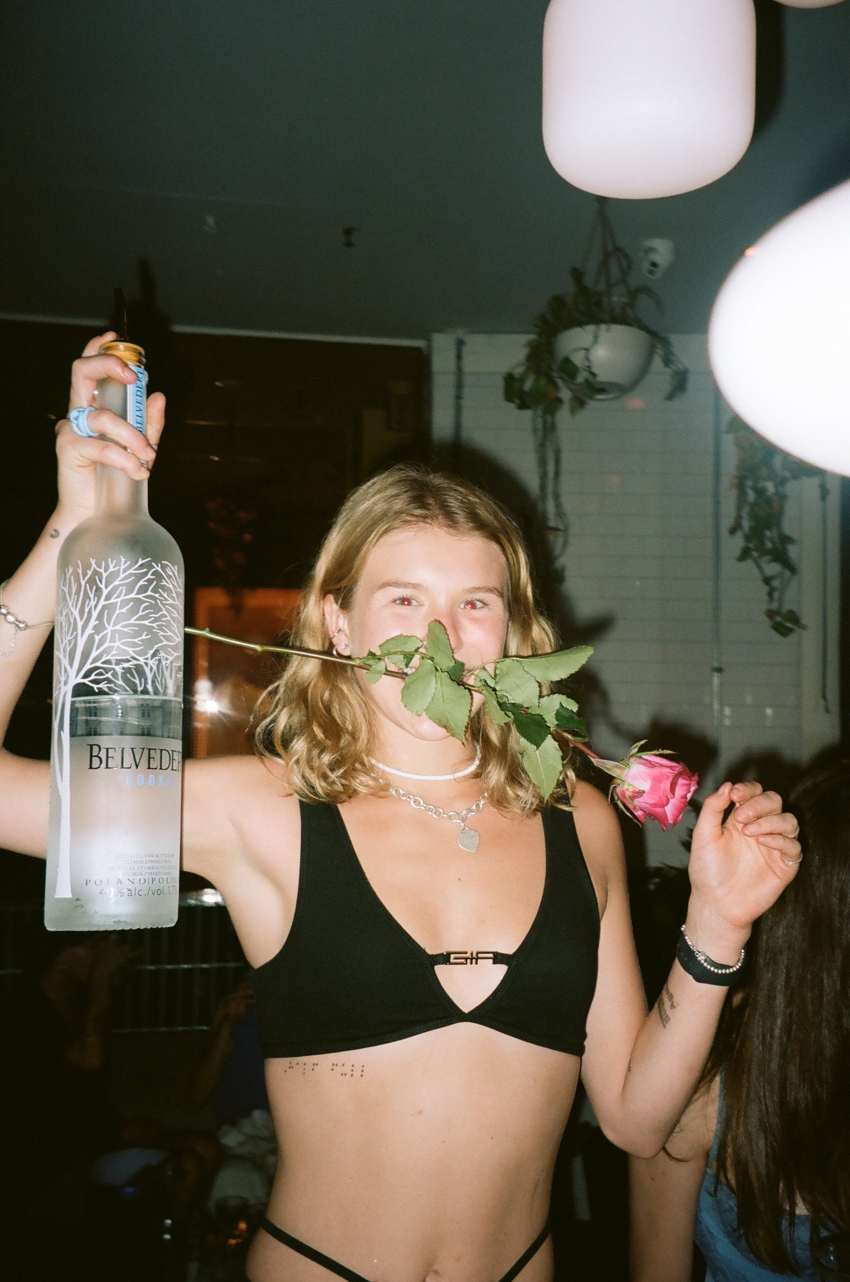 A young woman with blonde hair holding a pink rose and a bottle of Belvedere vodka, standing indoors at a party or celebration.