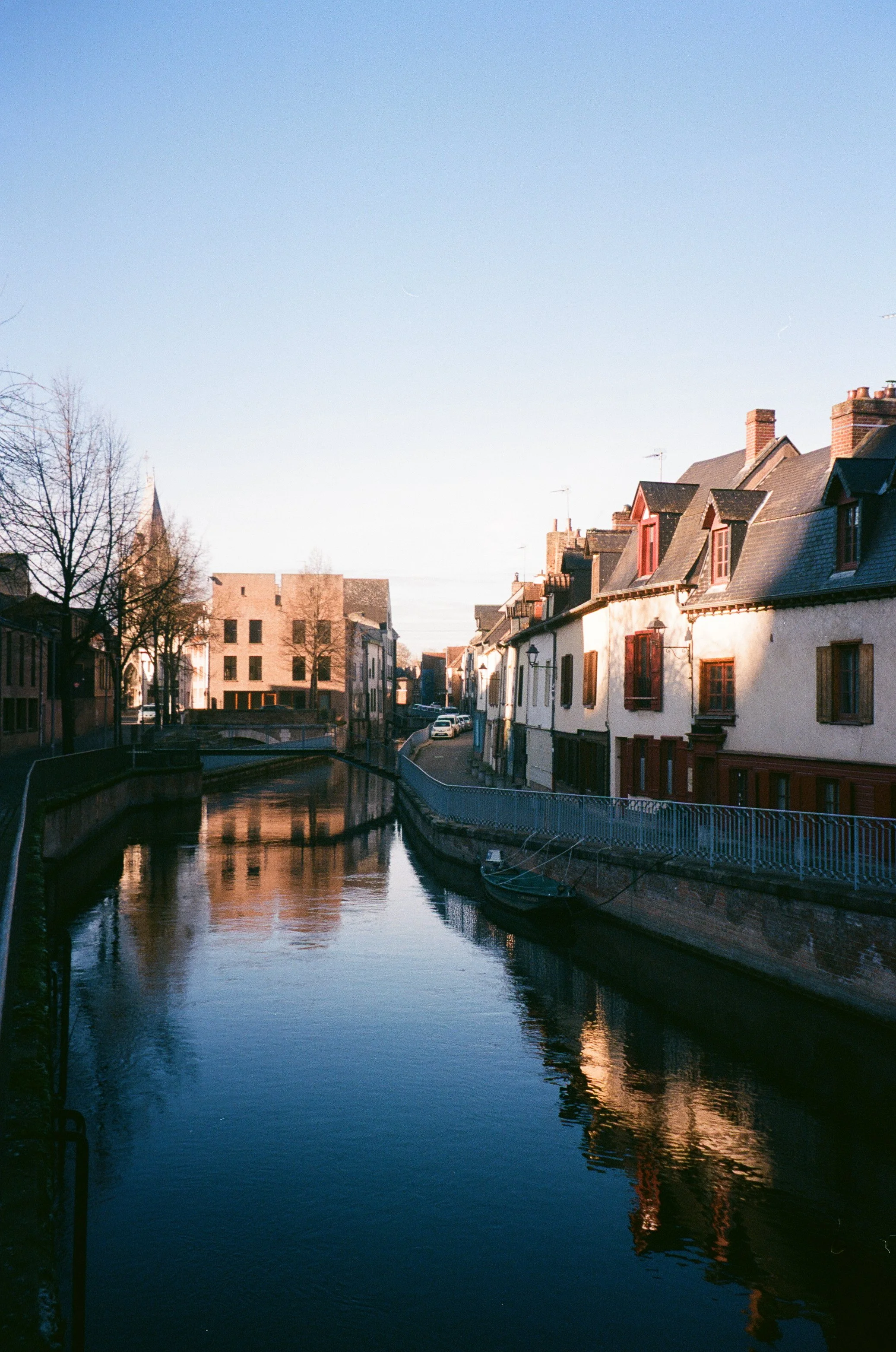 A peaceful canal scene with calm water reflecting the surrounding buildings and a clear sky, lined with houses and trees.
