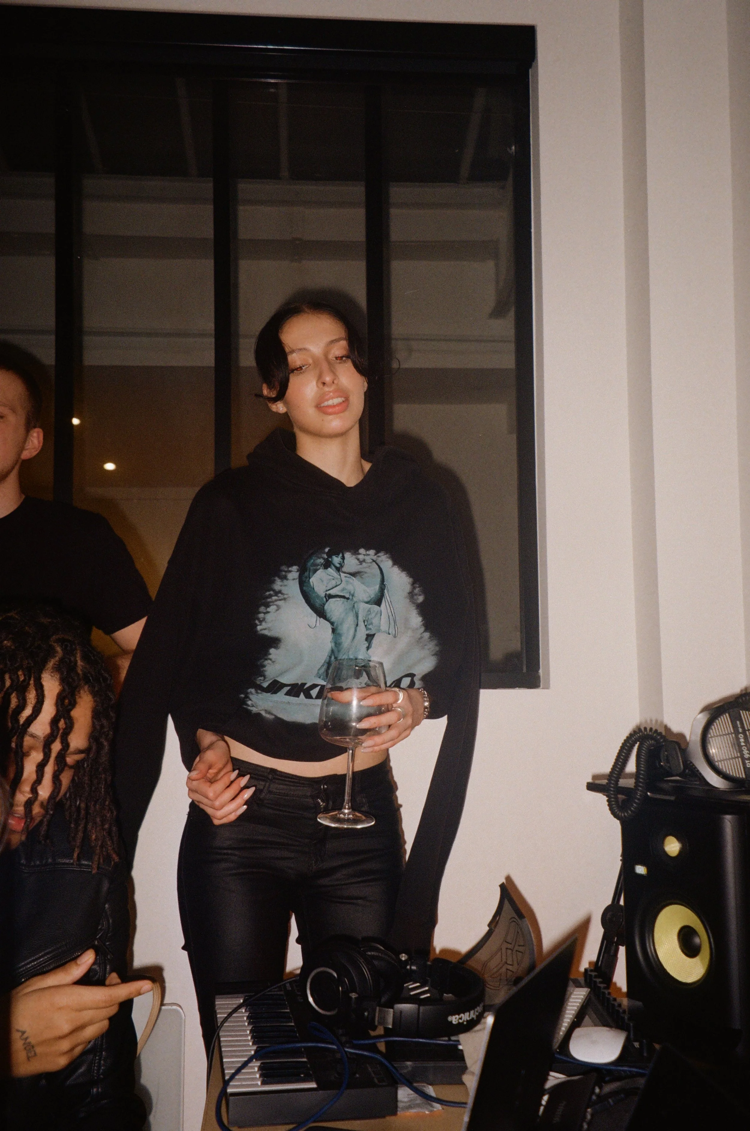 A young woman with short dark hair wearing a black graphic hoodie and black leather pants holding a glass of water, standing in a room with music equipment and a window behind her.