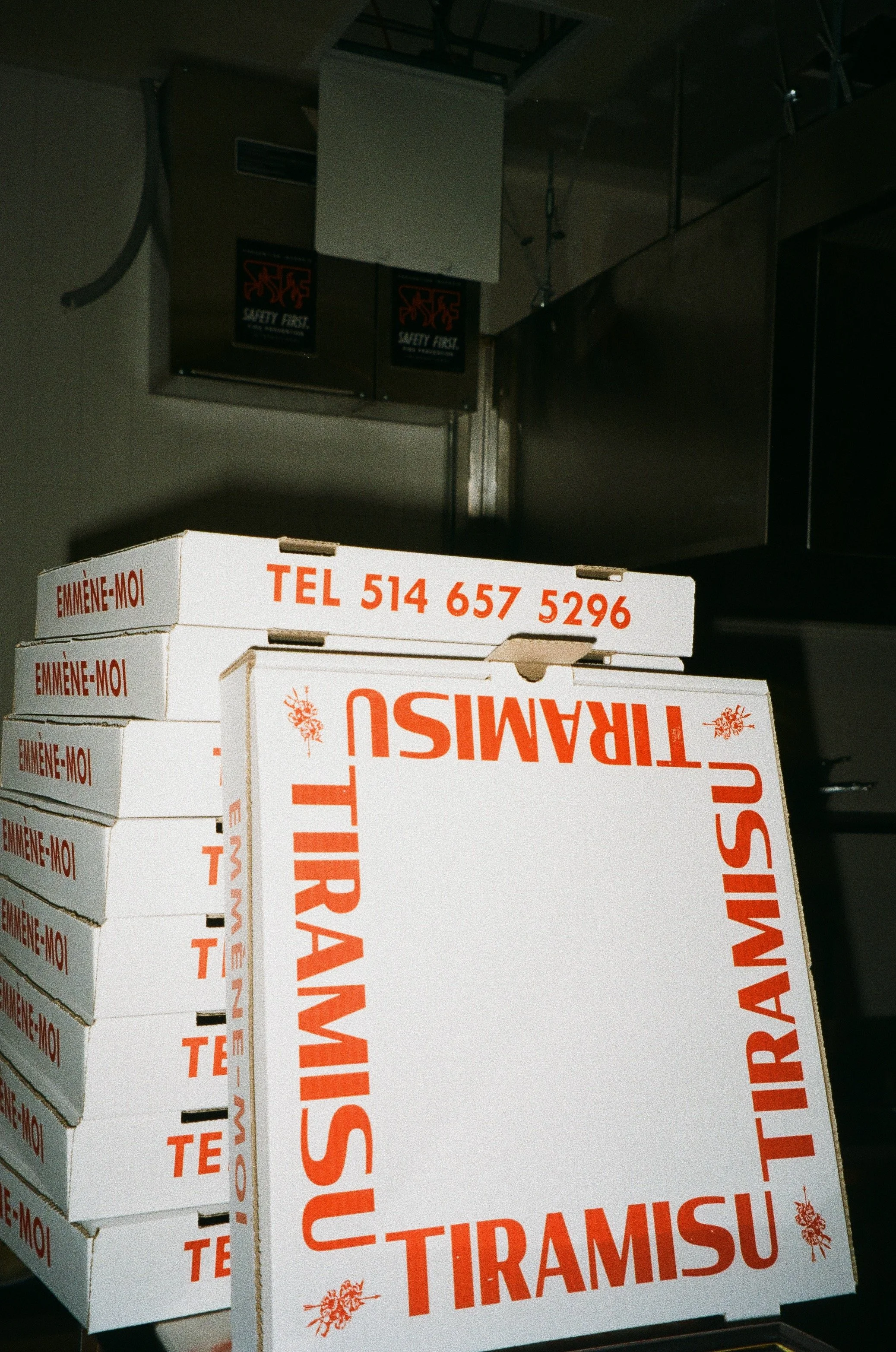 Stack of pizza boxes labeled 'TIRAMISU' and a phone number in red text, with some boxes stacked upright and others flat, in a kitchen or storage area.