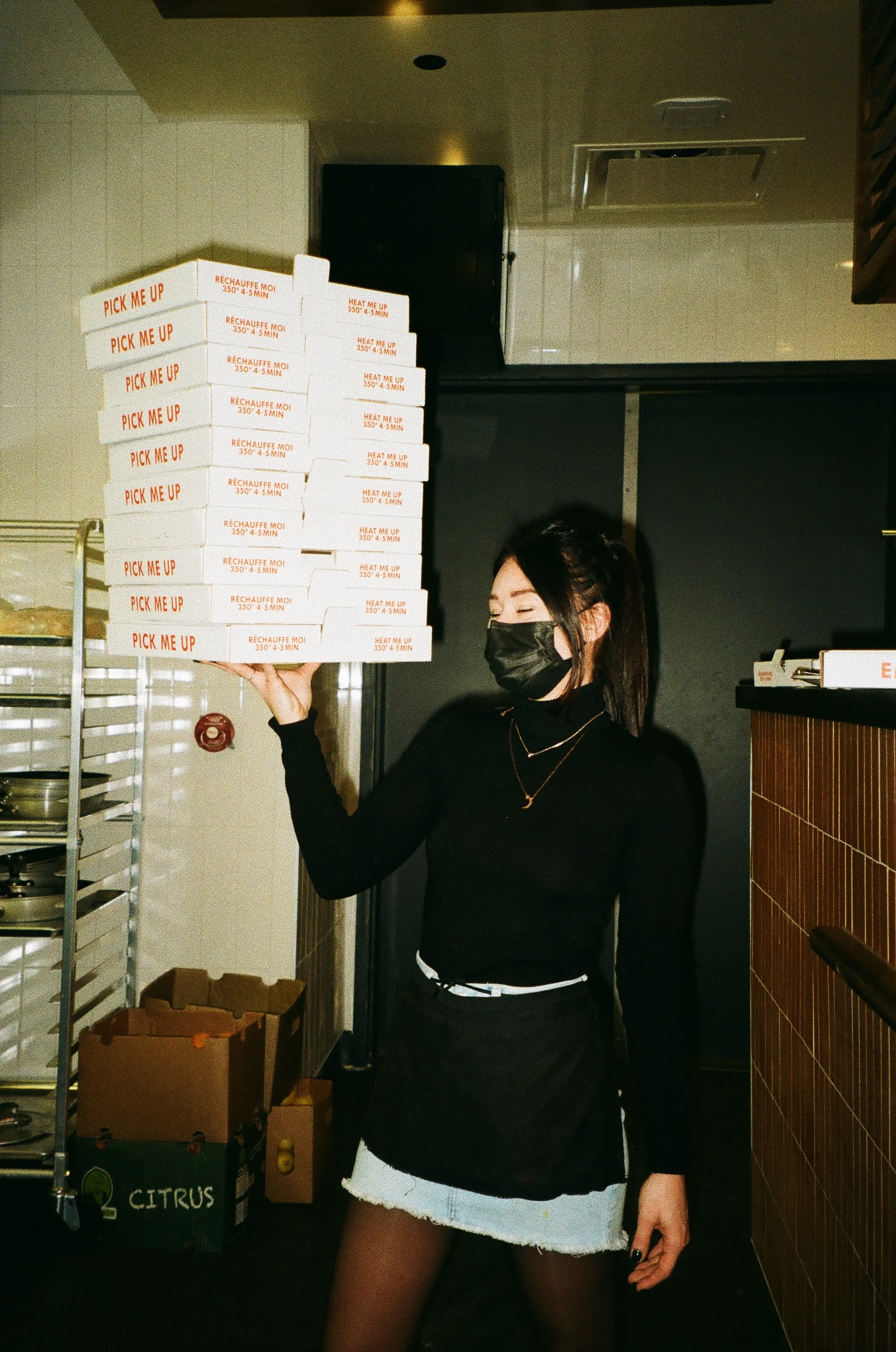 A woman wearing a black face mask, black sweater, and denim skirt is holding a stack of pizza boxes labeled "Pick Me Up" and "Heat Me Up" in a restaurant kitchen.