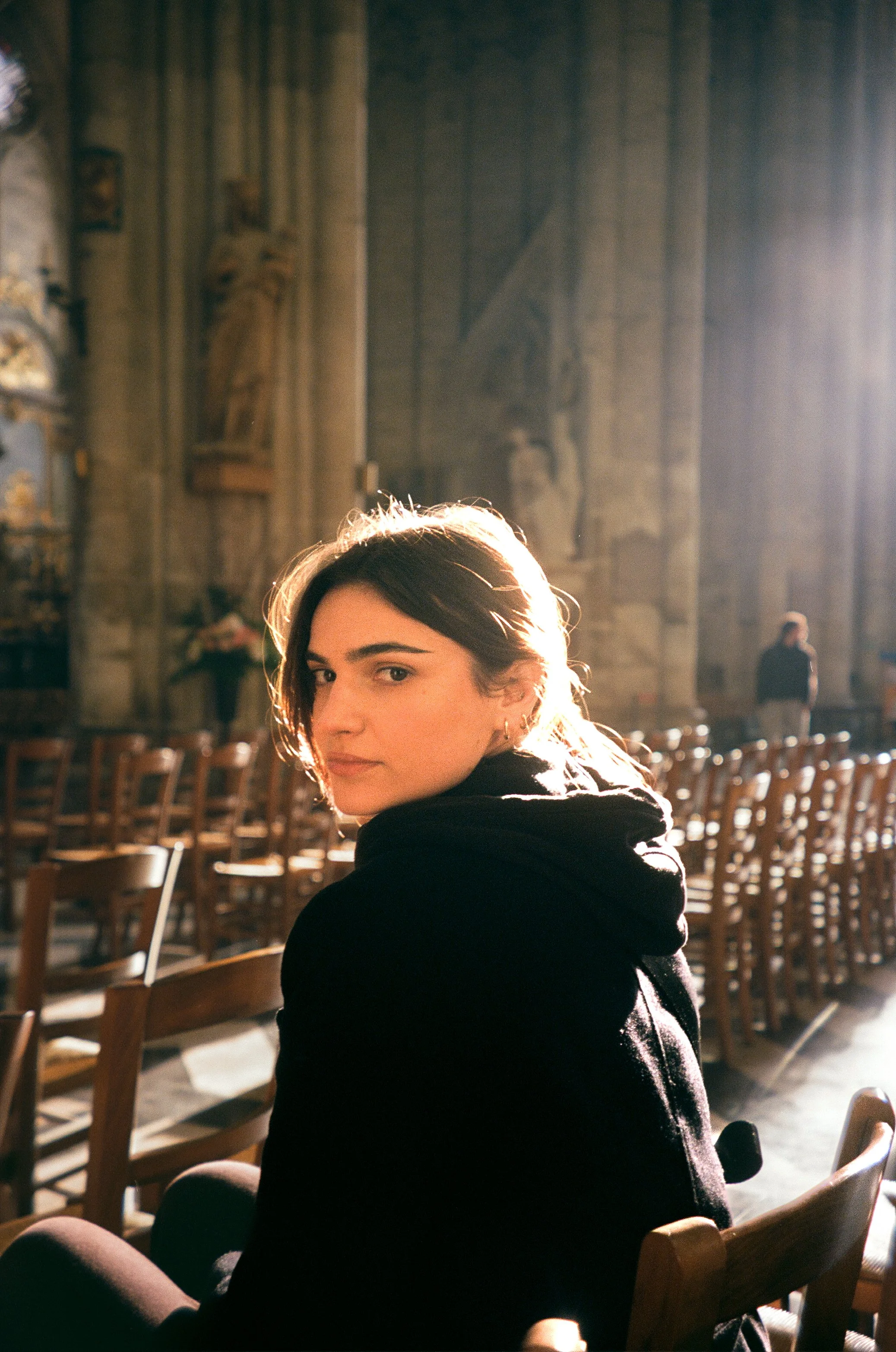 A young woman with dark hair, wearing a black hoodie, sitting on a chair inside a church, illuminated by sunlight filtering through windows, with empty chairs and religious statues in the background.