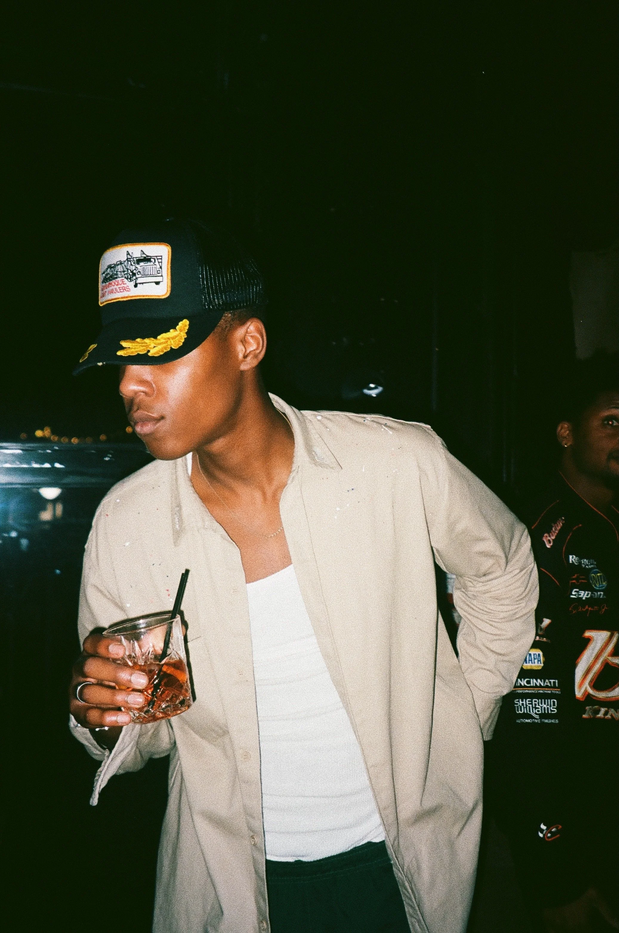 Young man wearing a black trucker cap with a patch and yellow embroidery, beige jacket, holding a glass of drink with ice and a straw, in a dimly lit setting.