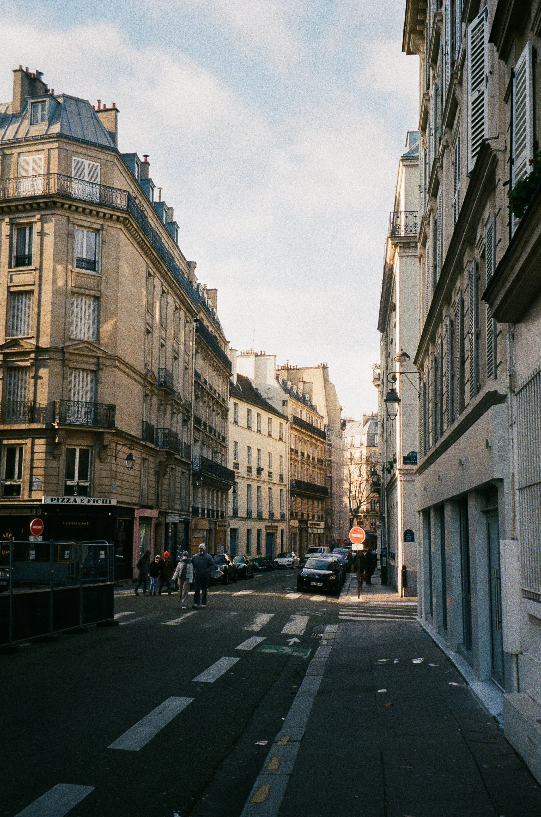 A street scene in a European city with multi-story historic buildings, pedestrians crossing, parked cars, and street signs.