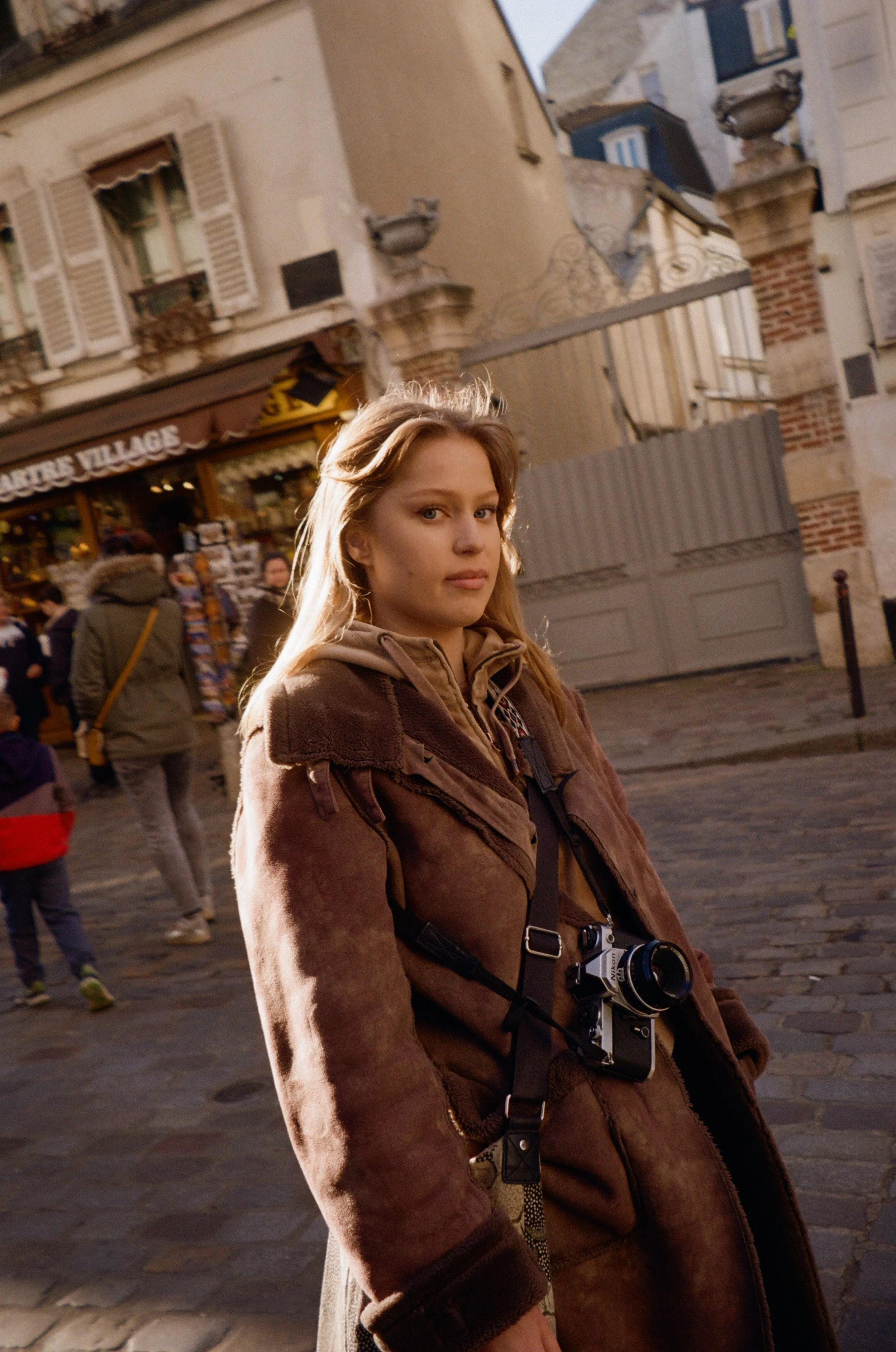 A young woman with long blonde hair standing on a cobblestone street, wearing a brown jacket, with a camera hanging from her neck, in an urban area with old buildings and other pedestrians in the background.