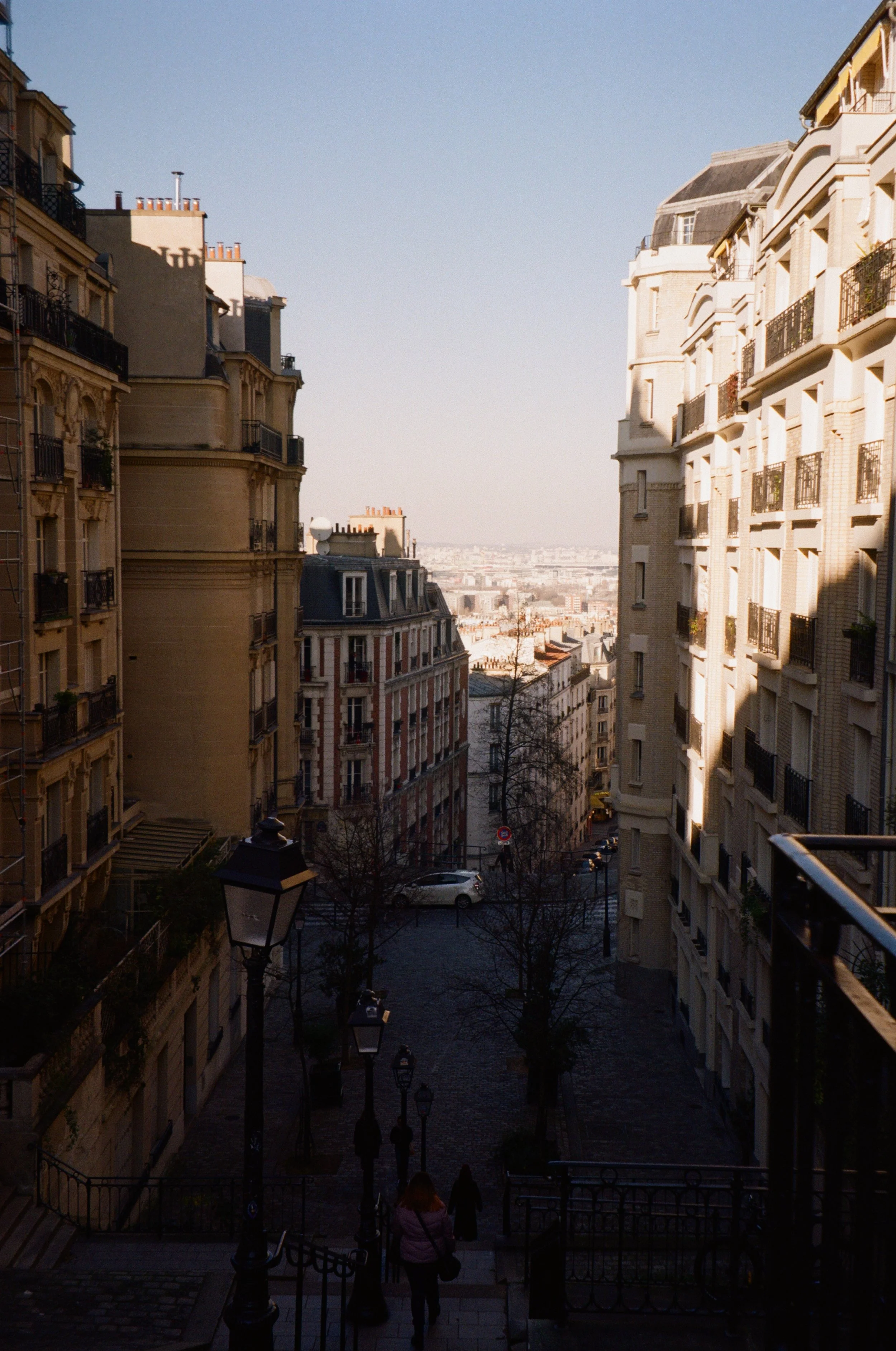 View of a European city street with tall apartment buildings, a cobblestone walkway, street lamps, and a few pedestrians, under a clear sky.