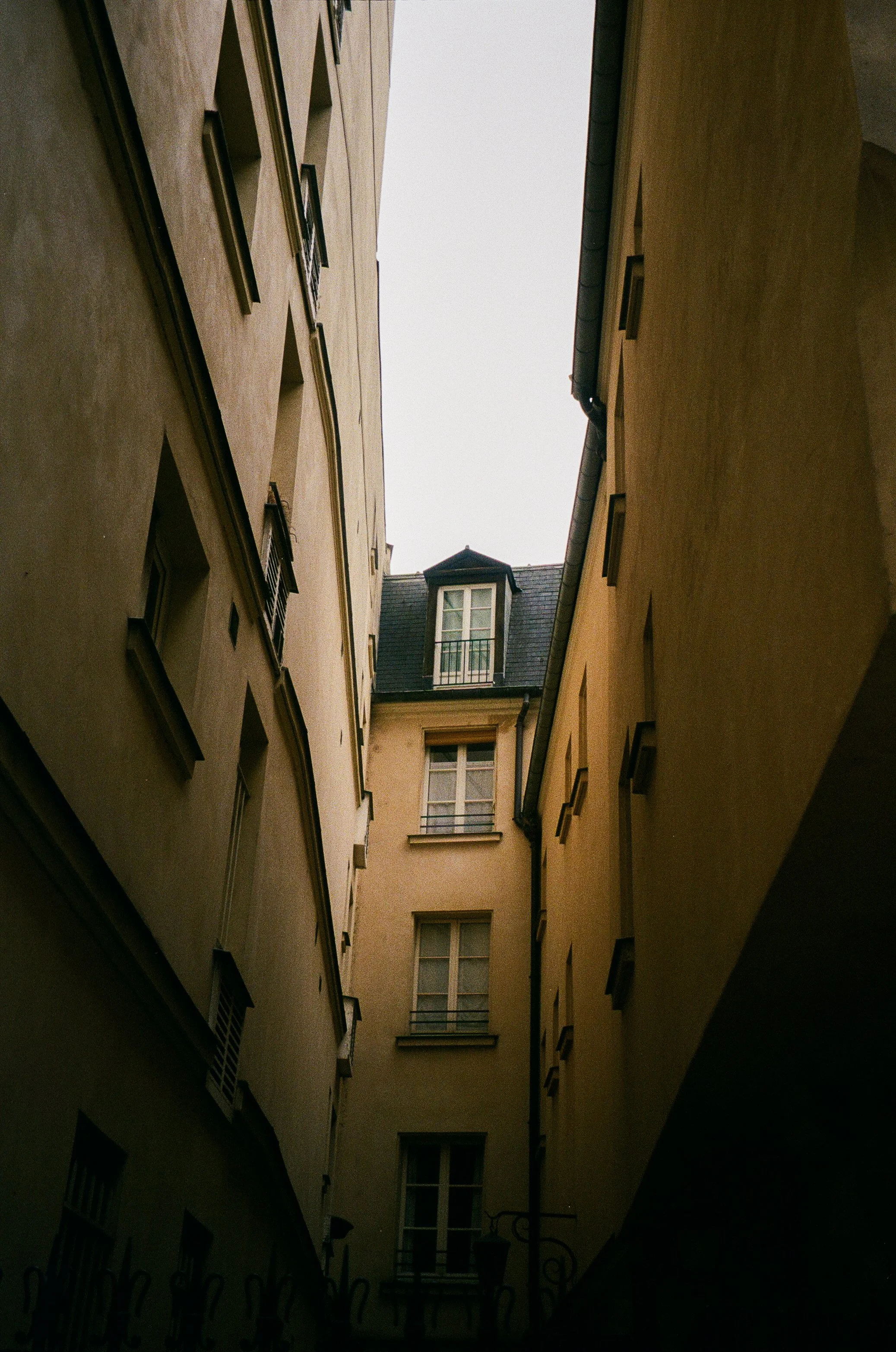 View of narrow courtyard between tall beige and yellow buildings with windows, leading up to a rooftop with dormer windows.