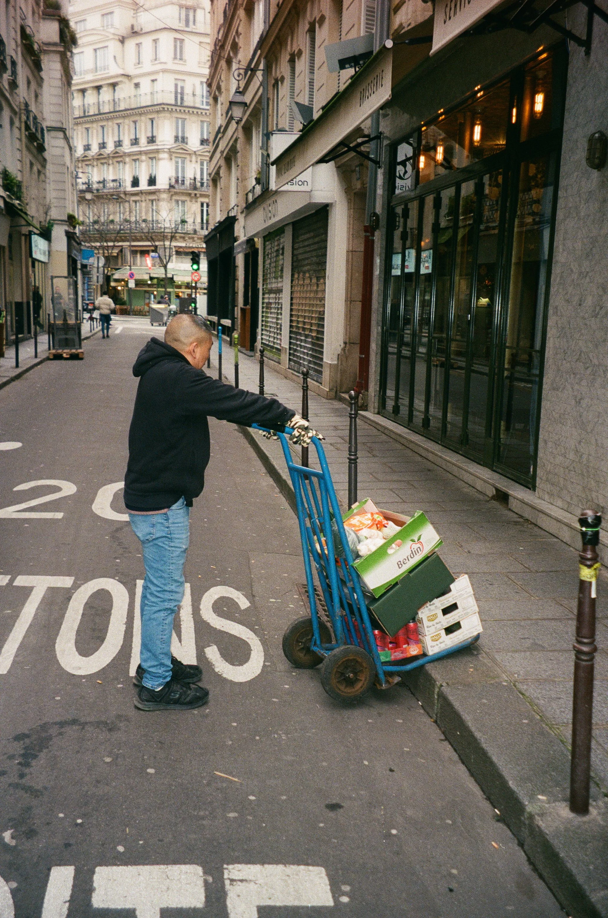 A man in a black hoodie and jeans pushing a blue hand truck loaded with boxes of groceries on a city sidewalk.