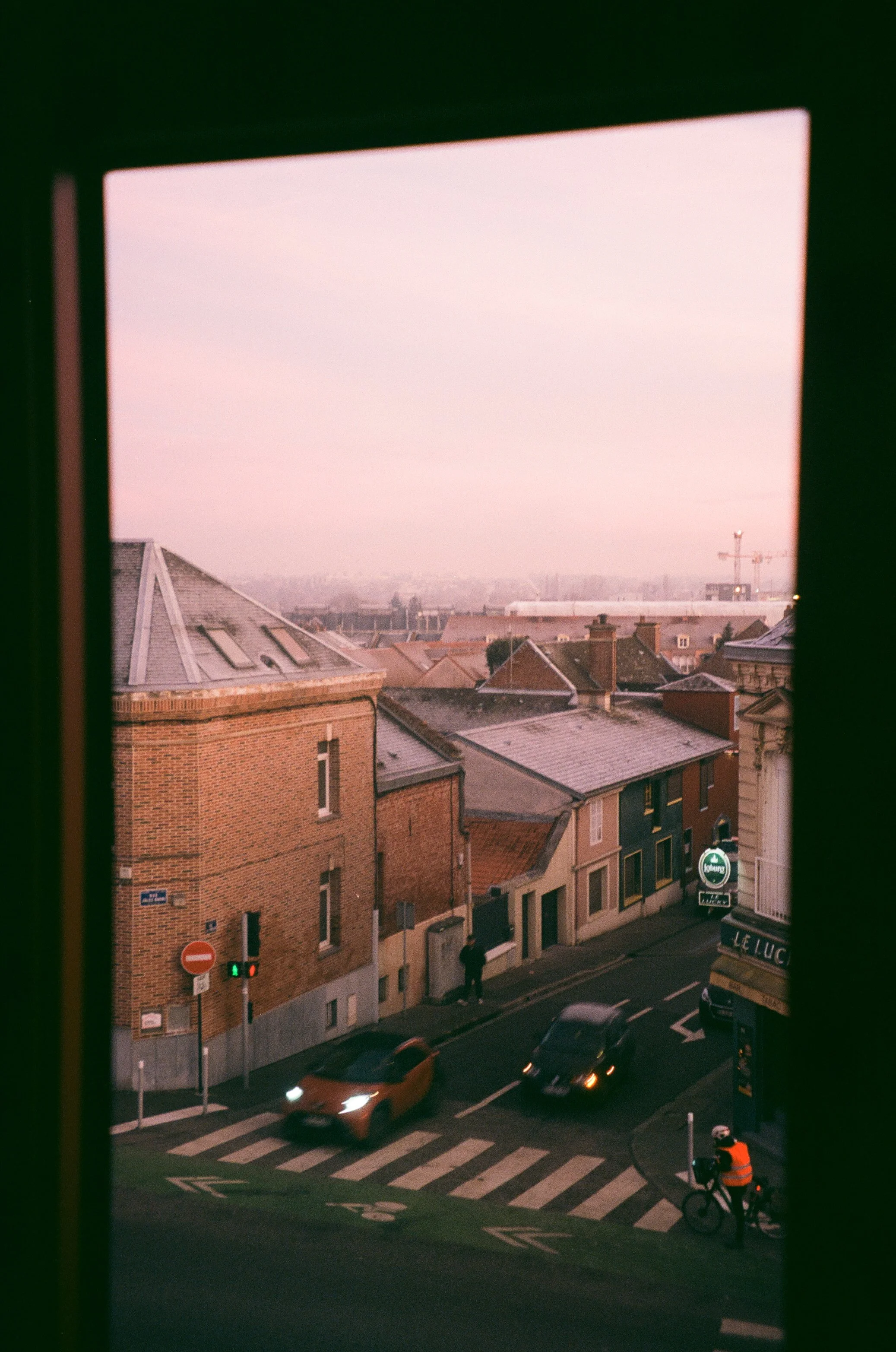 View through window showing a street with cars, a cyclist, and buildings, with a pink sky in the background.