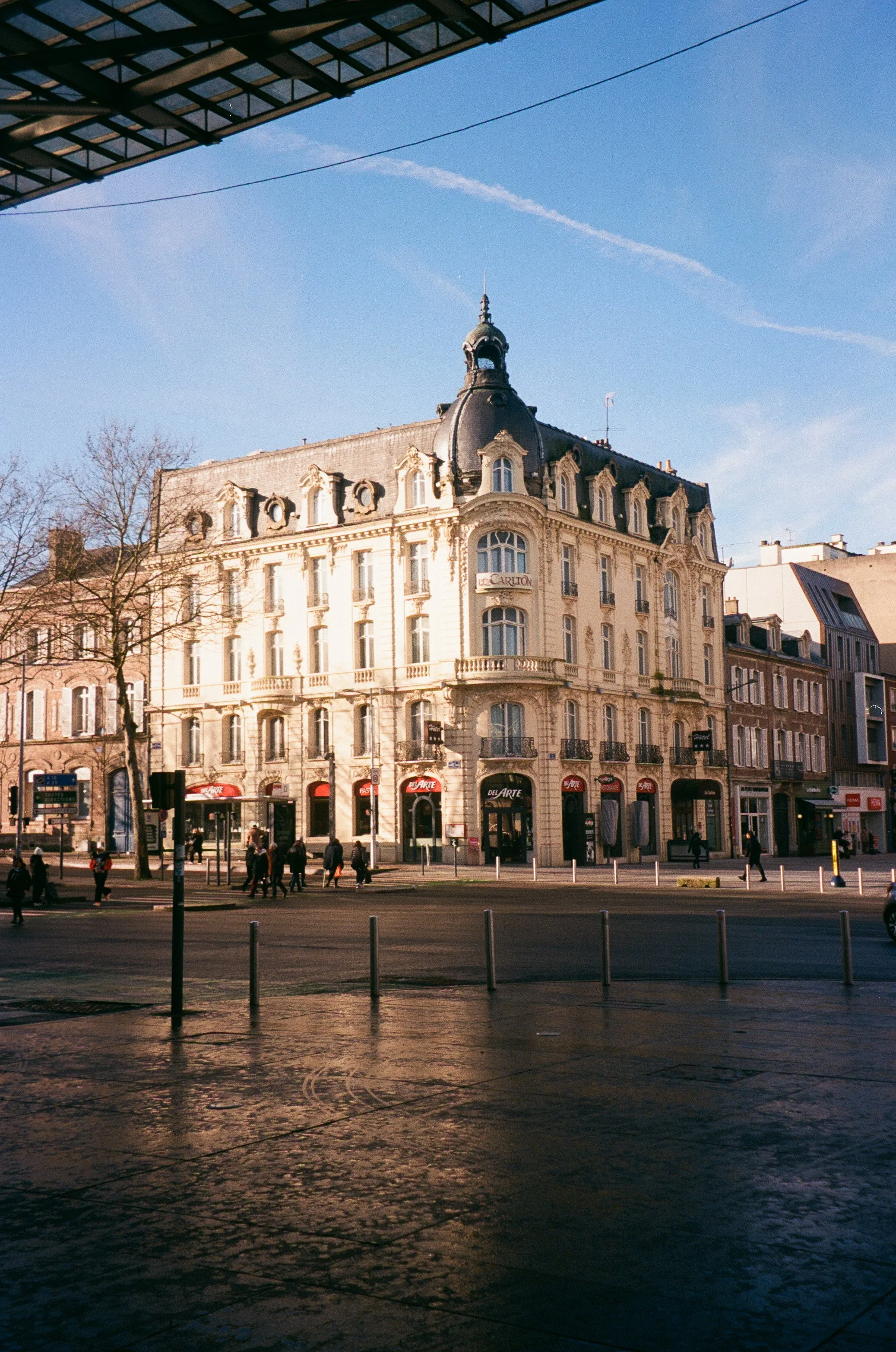 A historic European-style building with ornate architecture and curved balconies, situated on a bustling city street with pedestrians and shops, under a clear blue sky.