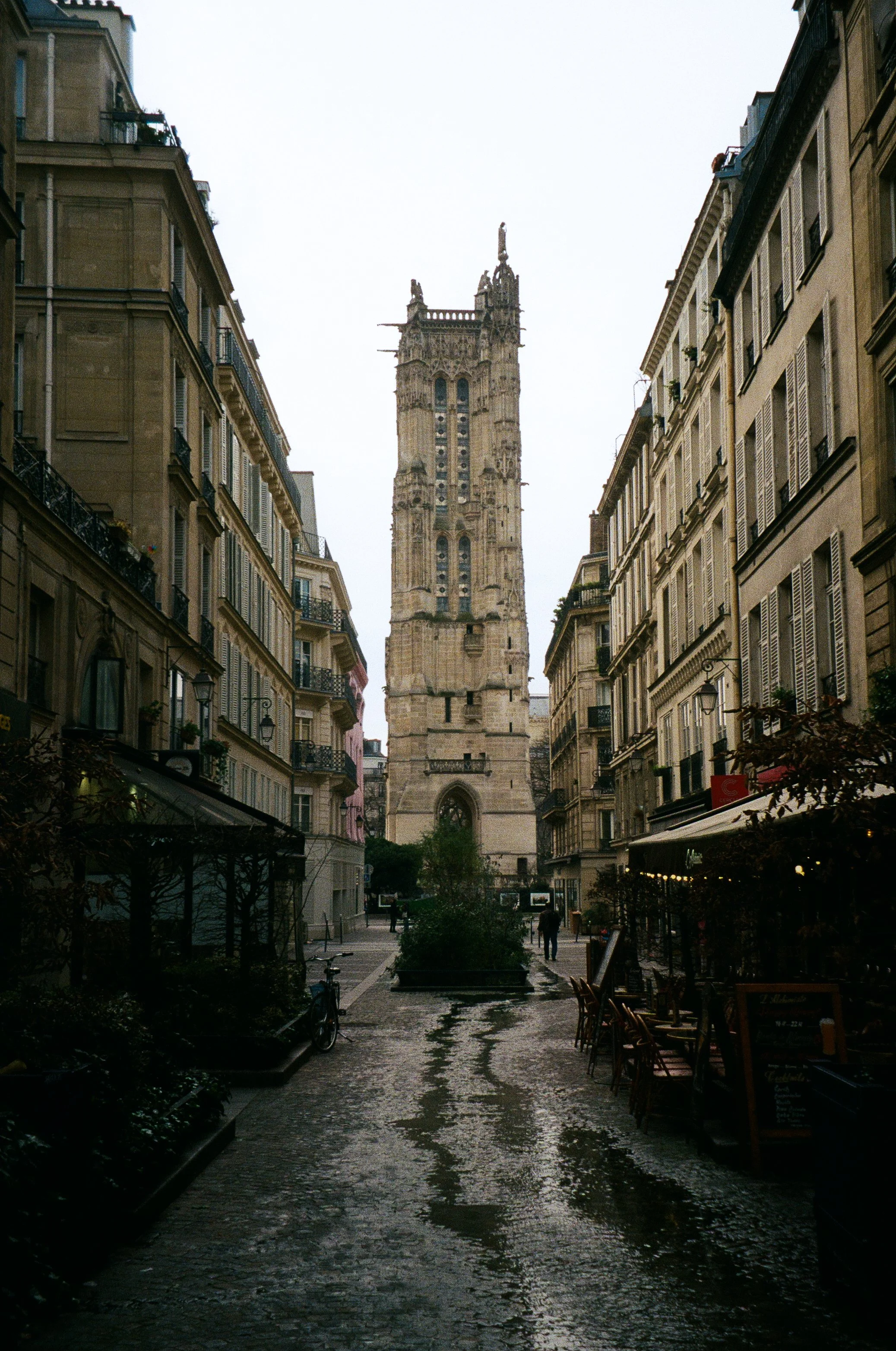 A view of a historic clock tower on a city street with cobblestone pavement, surrounded by buildings with balconies, some with plants, and outdoor cafes on the sides.