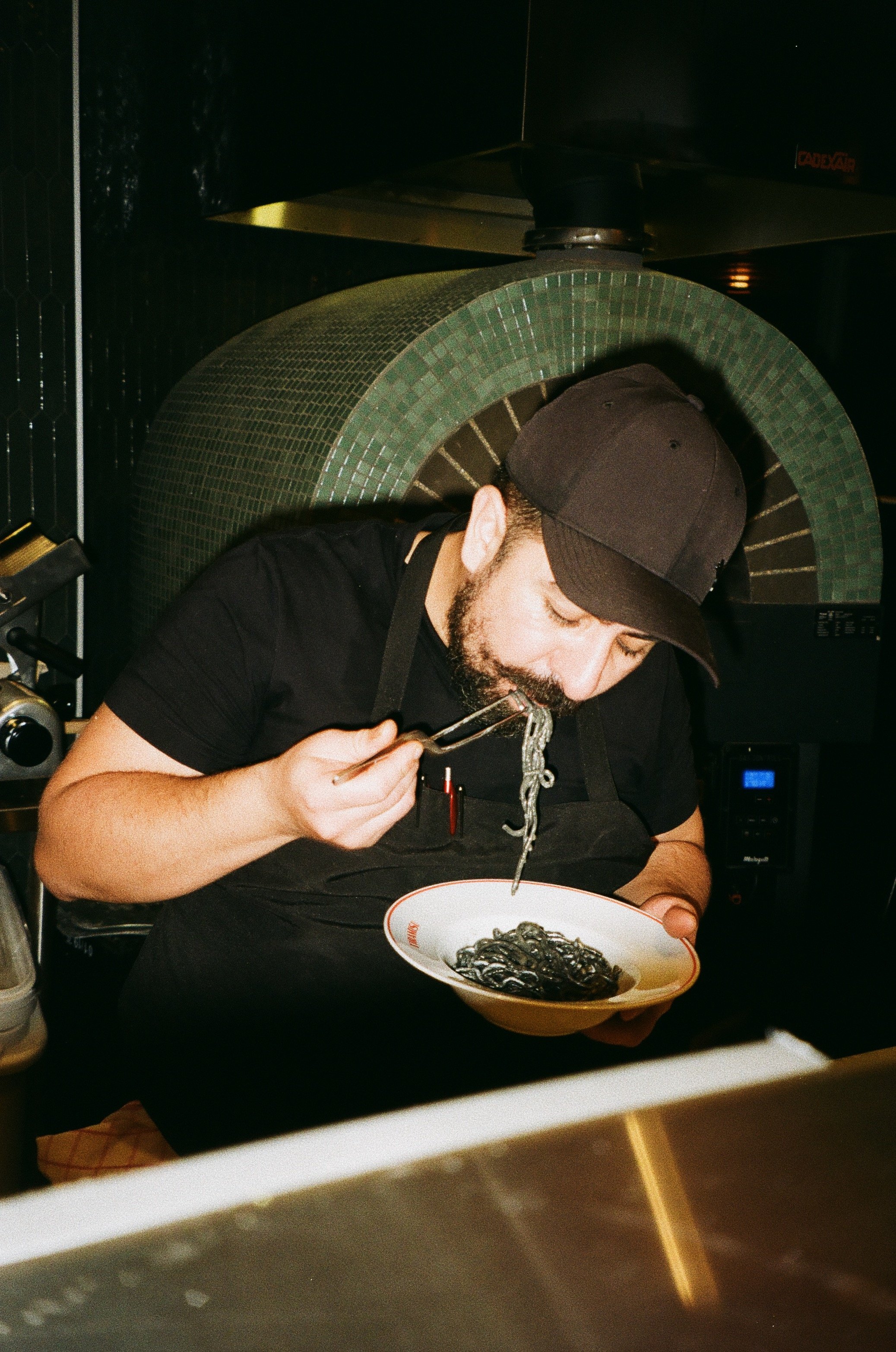 Man eating black pasta with tongs in a restaurant kitchen.